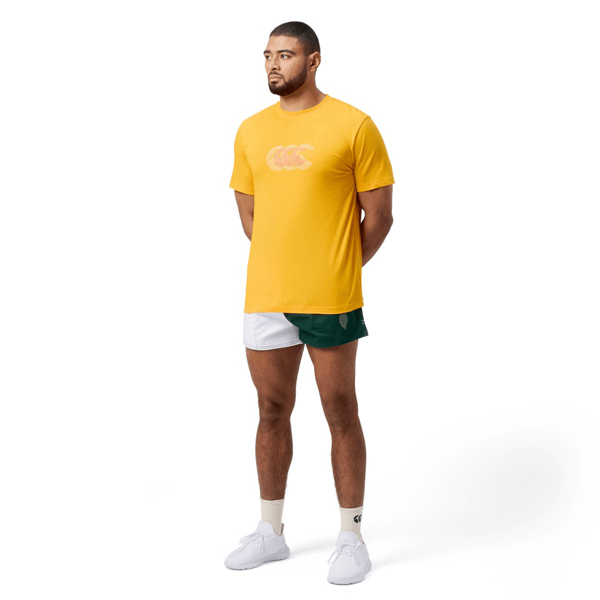 Someone in a classic fit yellow Canterbury Fundamental T-Shirt and white-green shorts stands sideward with arms behind their back. They wear white socks and shoes against a plain white background, showcasing the subtle Canterbury CCC logo design.