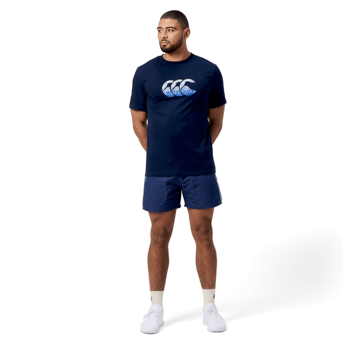 A man in a Canterbury Fundamental T-Shirt with a classic fit and CCC logo stands against a white background. He pairs it with blue shorts, his hands behind his back, wearing white socks and sneakers.