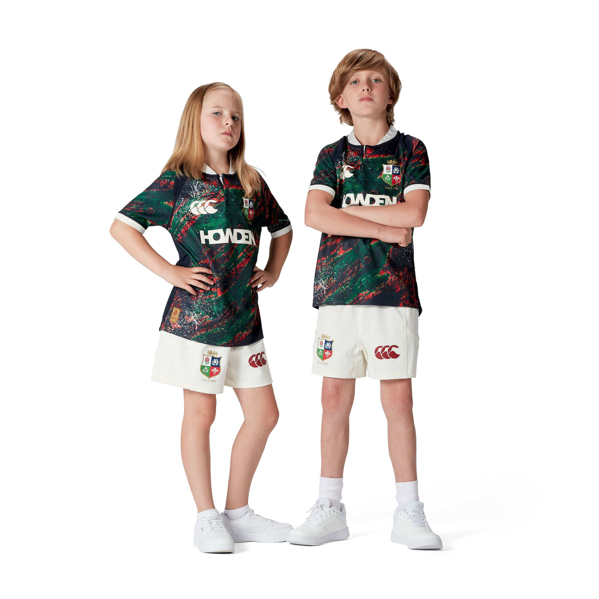 Two kids stand confidently in matching sports uniforms with a green and red abstract pattern and white shorts. Made from moisture-wicking fabric, these outfits echo the style of the Youth British & Irish Lions 2025 Warm Up Jersey by Canterbury against a clean, white backdrop.