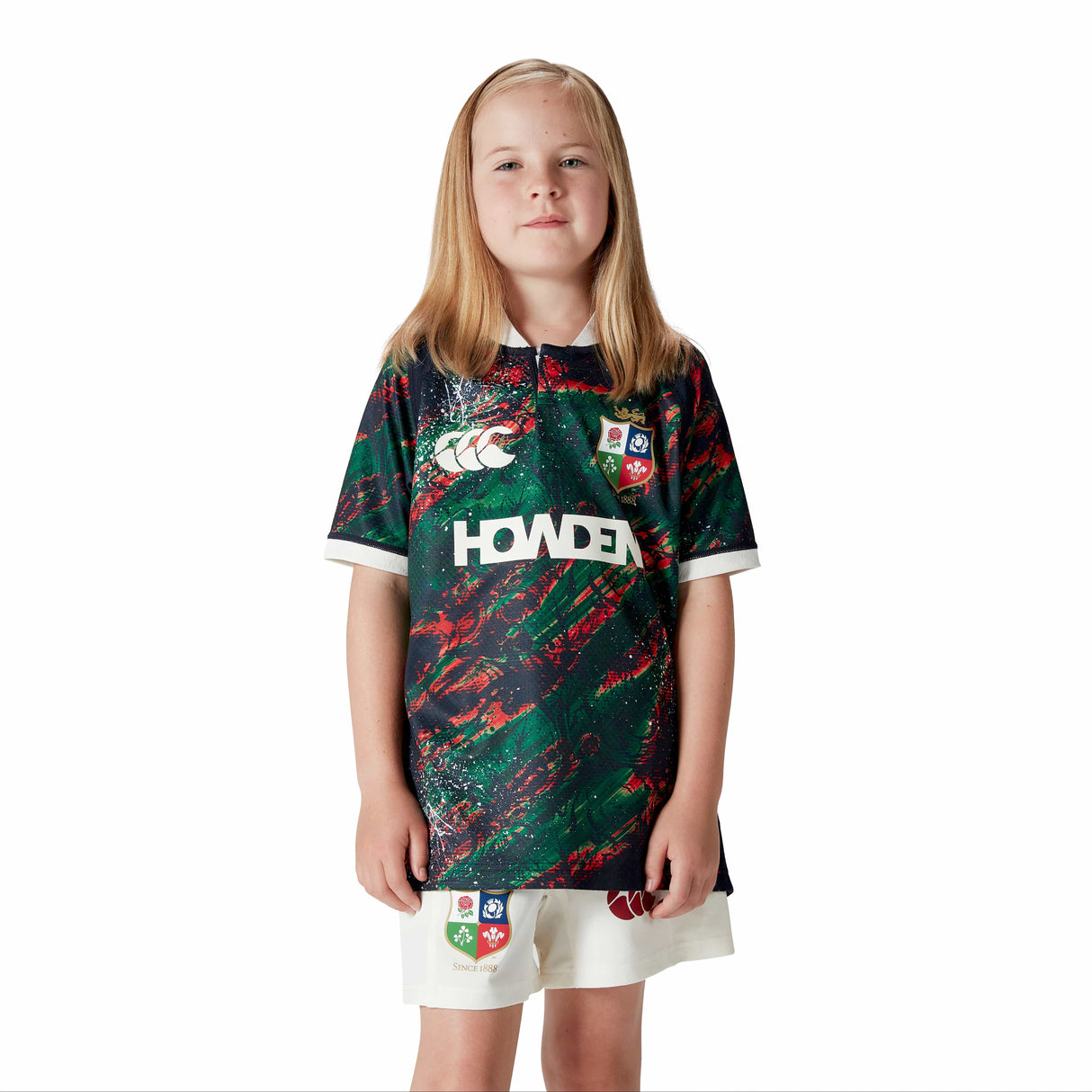 A child wearing the Canterbury Youth British & Irish Lions 2025 Warm Up Jersey and white shorts poses against a plain white background, highlighting its vibrant design.