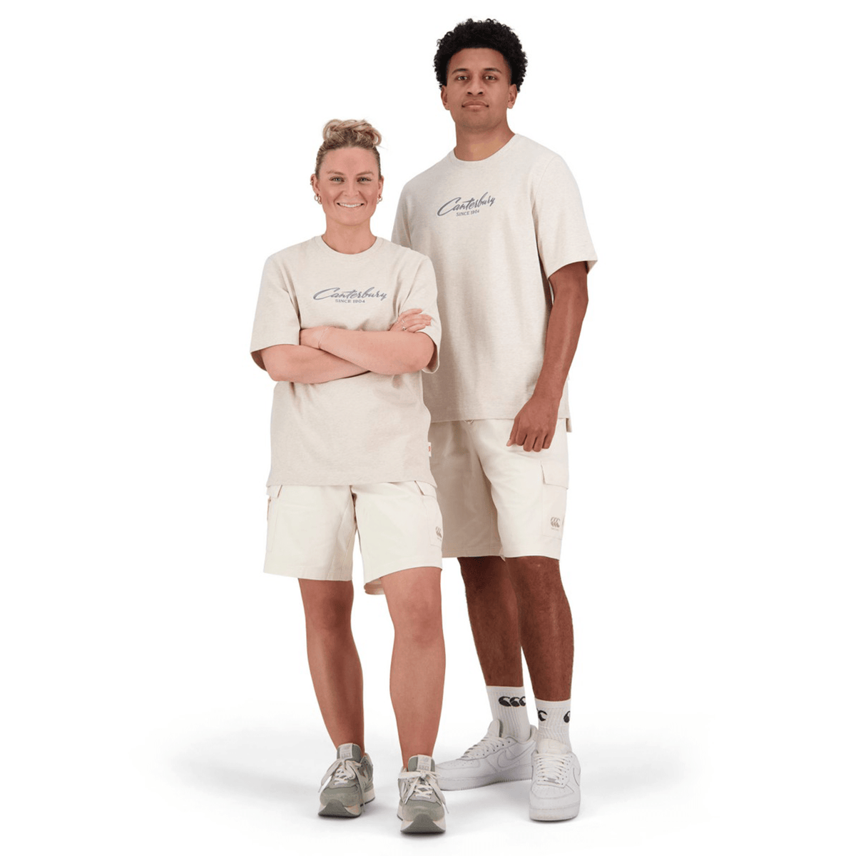 Two people stand smiling at the camera, dressed in matching Canterbury Heritage Script T-Shirts made from organic cotton. Their coordinated light beige outfits feature shorts and t-shirts, all set against a plain white background.