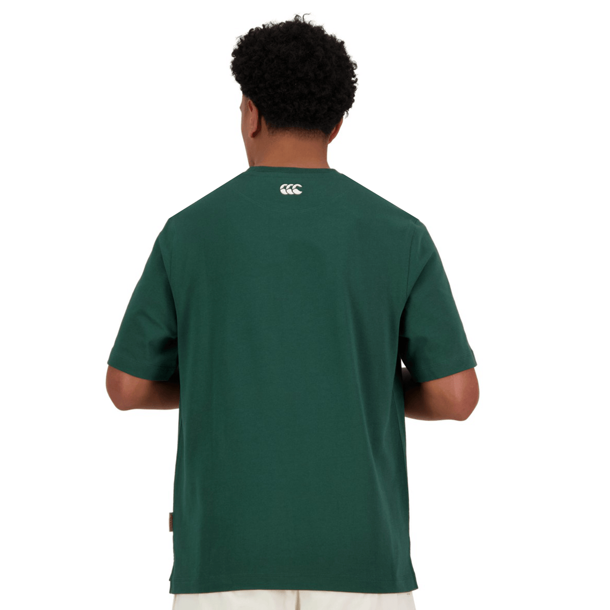 A person with curly hair faces away, wearing a dark green Canterbury Heritage CCC T-Shirt crafted from organic cotton, showcasing a small logo on the back. It pairs perfectly with light-colored pants, embodying Canterbury of New Zealand's timeless style.