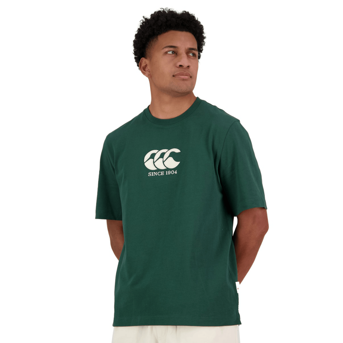 A person wears a Canterbury Heritage CCC T-Shirt in dark green, featuring the iconic logo and "Since 1904" text. Crafted from organic cotton, this stylish piece by Canterbury celebrates timeless design and sustainability, with their arms behind their back.