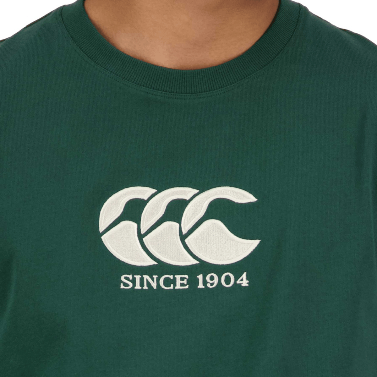 Close-up of a person wearing the green Canterbury Heritage CCC T-Shirt, showcasing Canterbury's iconic white logo with three "C"s and "SINCE 1904" embroidered below. Crafted from organic cotton, it blends comfort with timeless tradition.