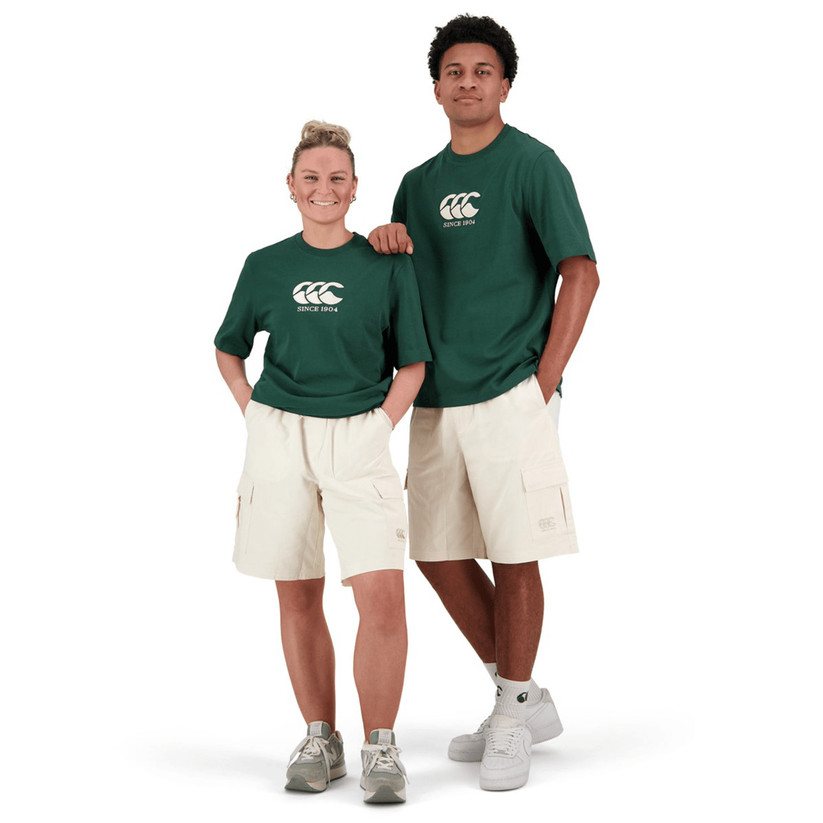 Two individuals stand side by side, each wearing a green Canterbury Heritage CCC T-Shirt with white logos and beige shorts made from organic cotton.