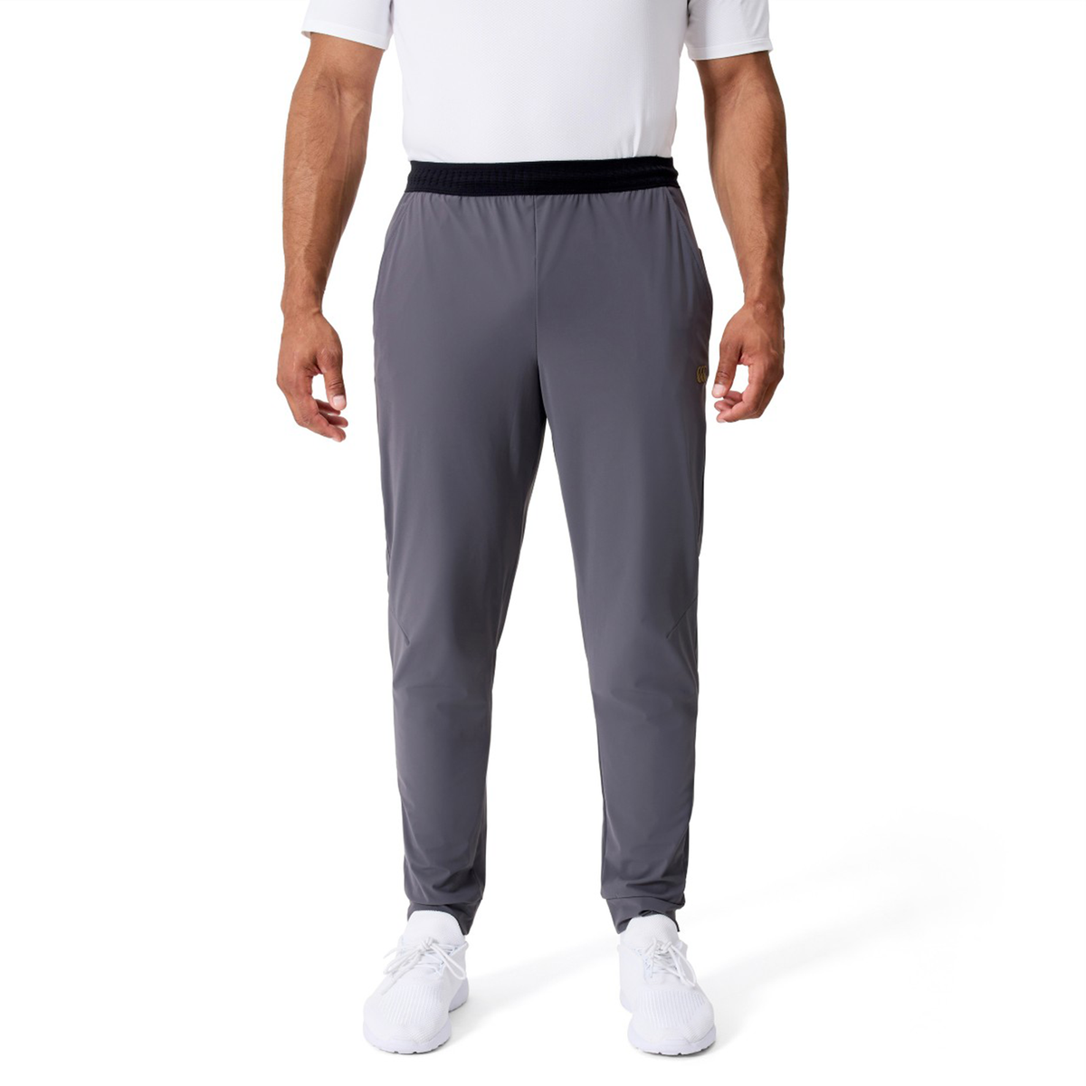 A man stands facing forward against a plain white background wearing a white short-sleeve shirt, Canterbury Everest Woven Pants by Canterbury in gray, and white sneakers.