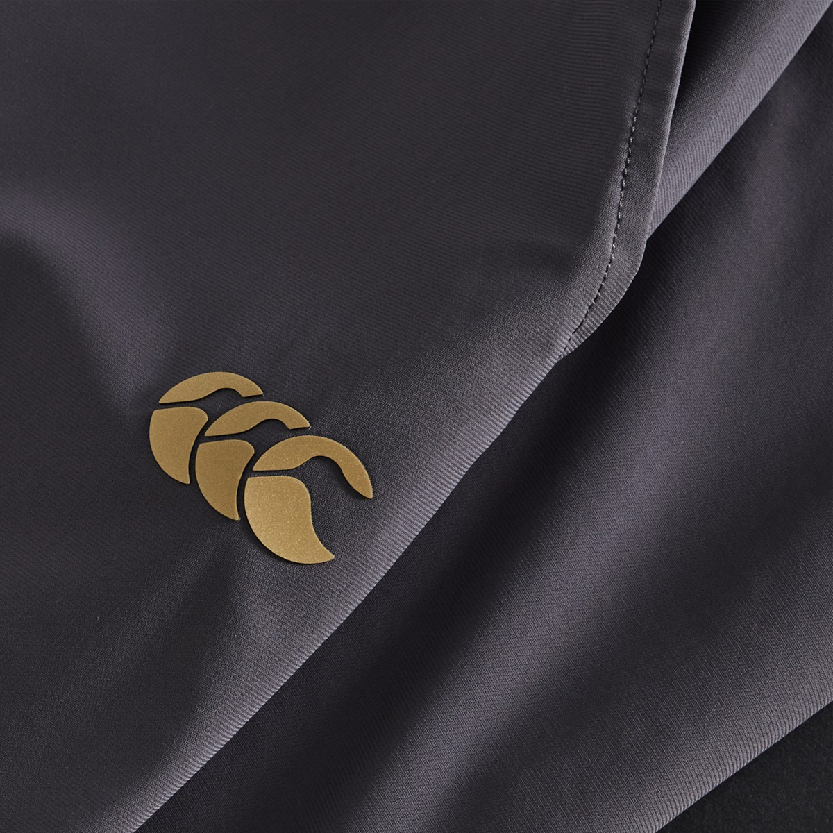 Close-up of Canterbury Everest Woven Pants in dark gray, featuring a gold Canterbury logo on the lower left—ideal for rugby or performance training.