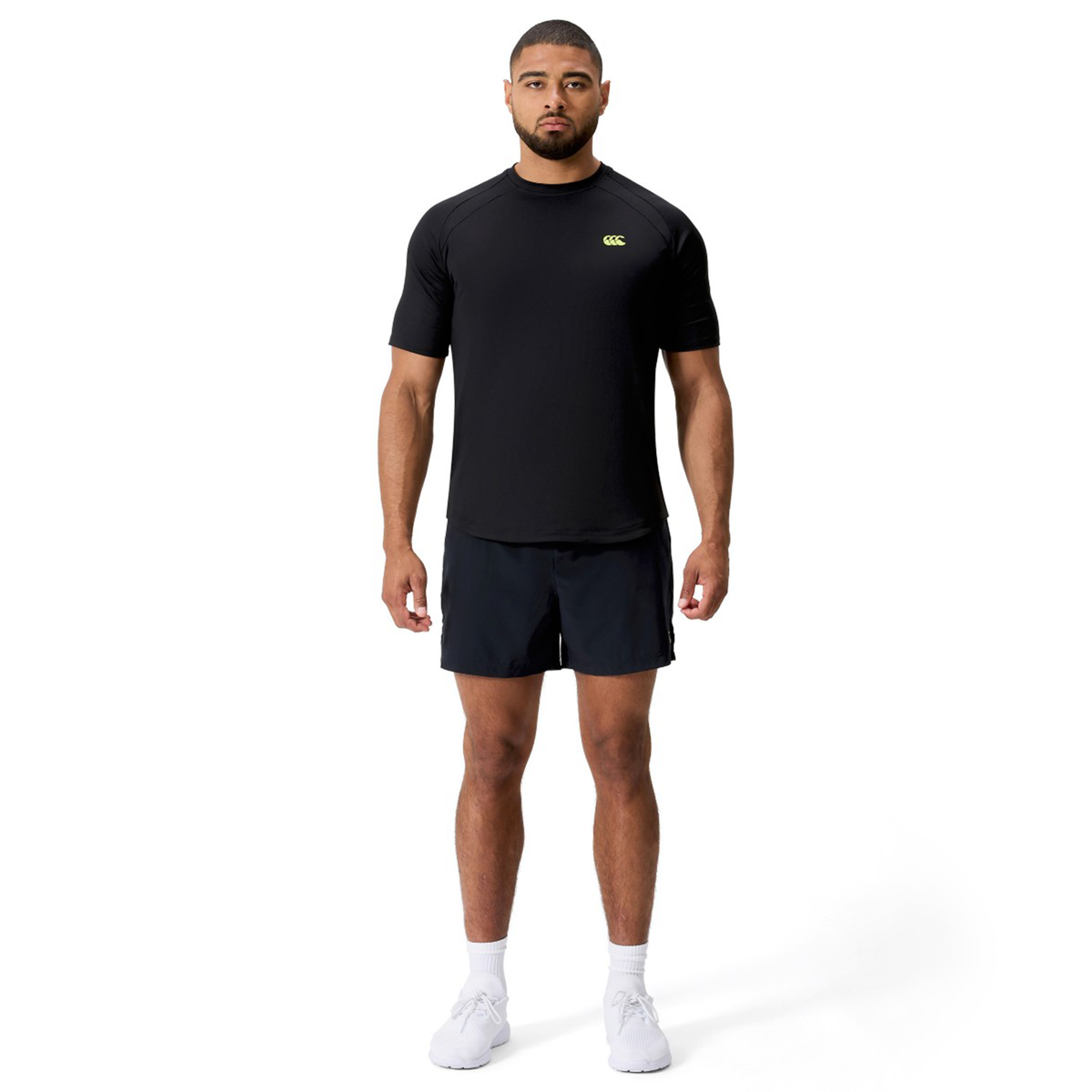 A man faces forward against a white background, wearing the Canterbury Everest Training Tee in black, paired with black shorts and white athletic shoes.