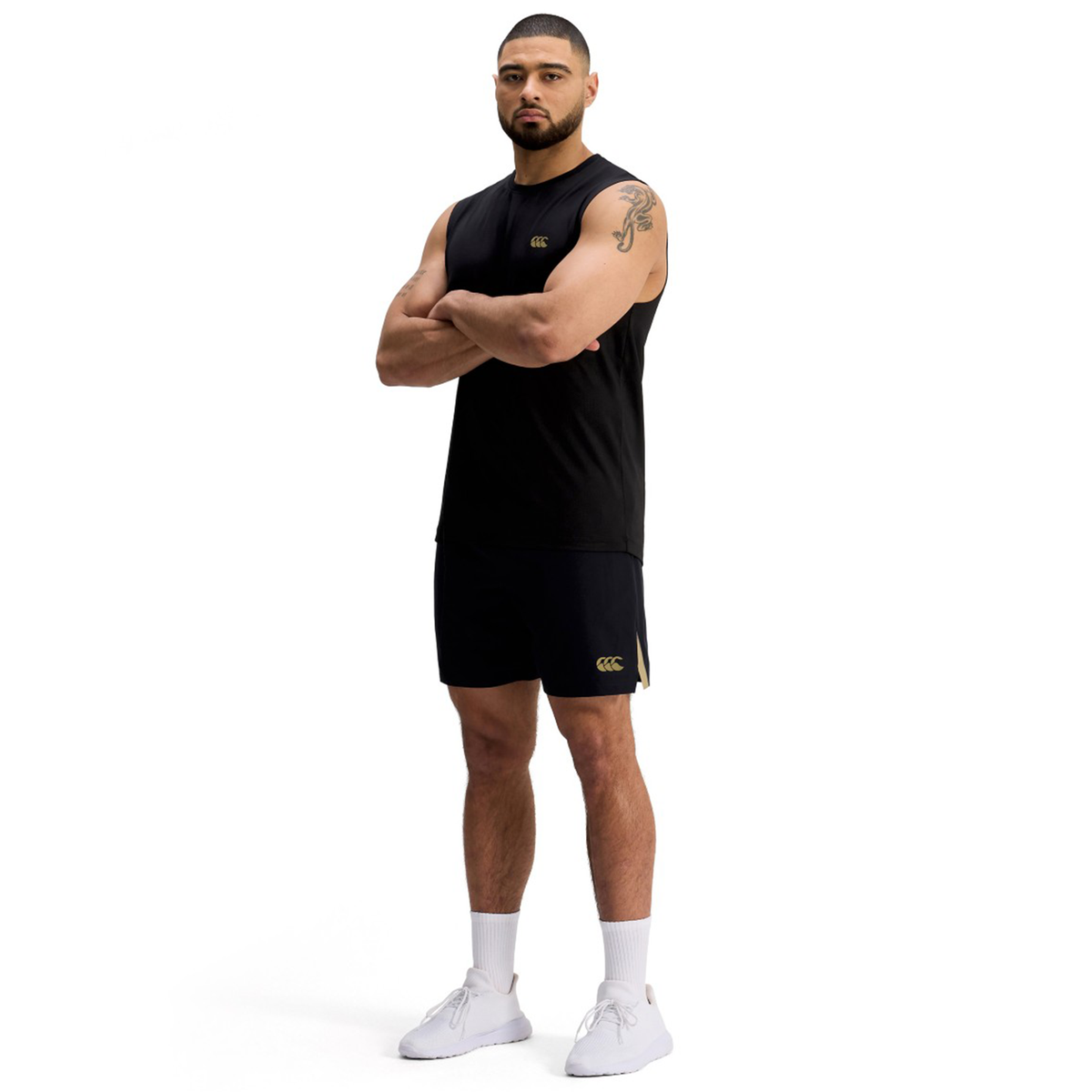 A man stands with arms crossed, wearing black Canterbury Tempo+ 7" Training Shorts, a black sleeveless shirt, white socks, and white athletic shoes against a plain white background.