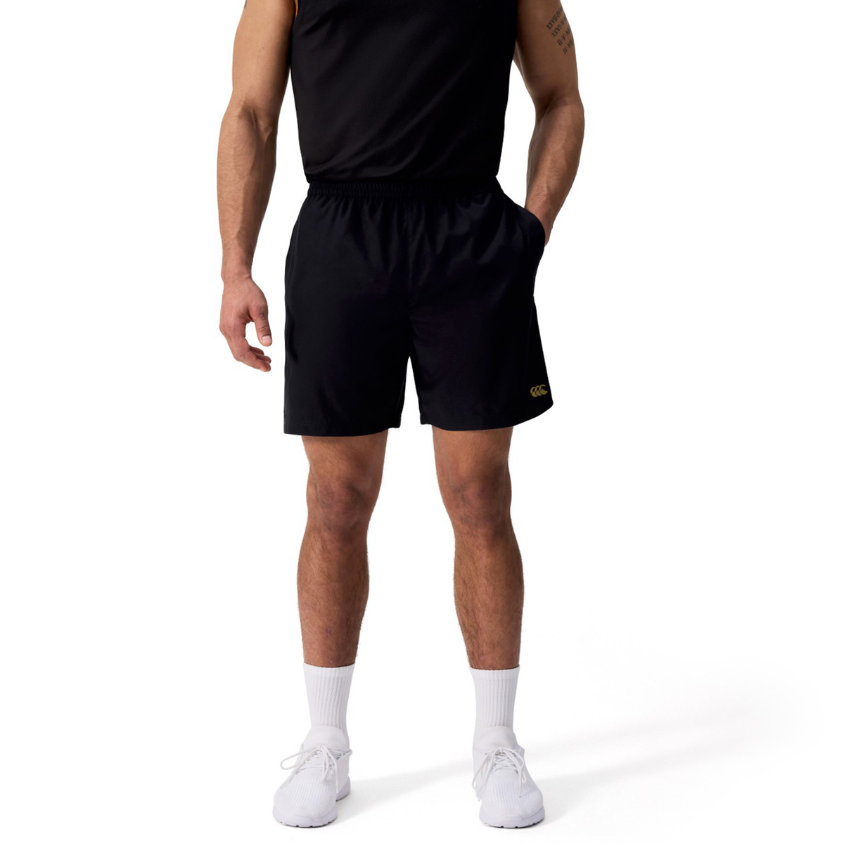 A person wears black Canterbury Tempo+ 7" Training Shorts, an athletic shirt, white socks, and white sneakers, standing with hands in pockets against a plain background.