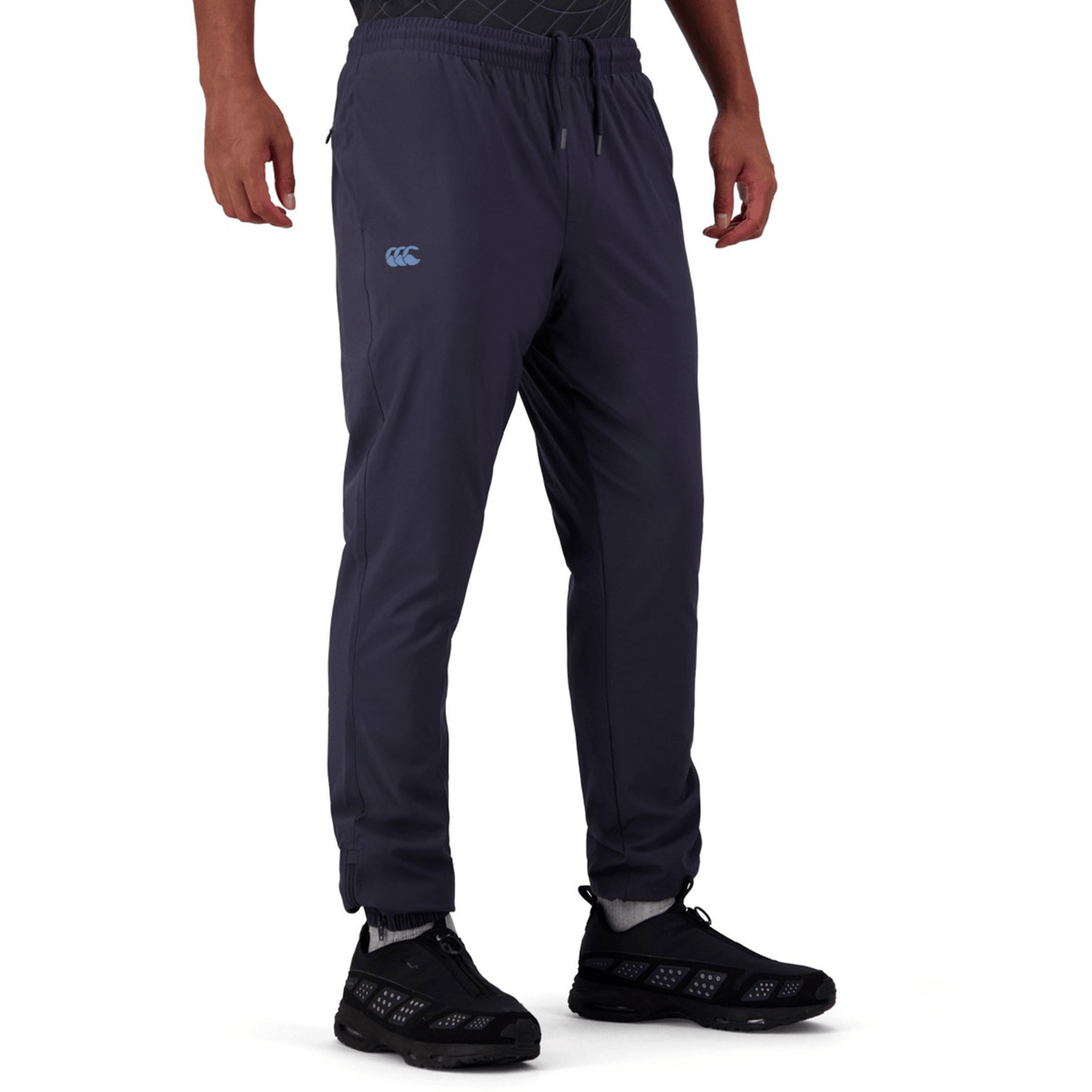 A person sports Canterbury Tempo+ Training Trackpants, featuring a tapered fit with a light blue logo on the thigh. The dark joggers, crafted from bi-stretch fabric for flexibility, are paired with black sneakers. Hands rest relaxed by the sides while the upper body remains elusive.