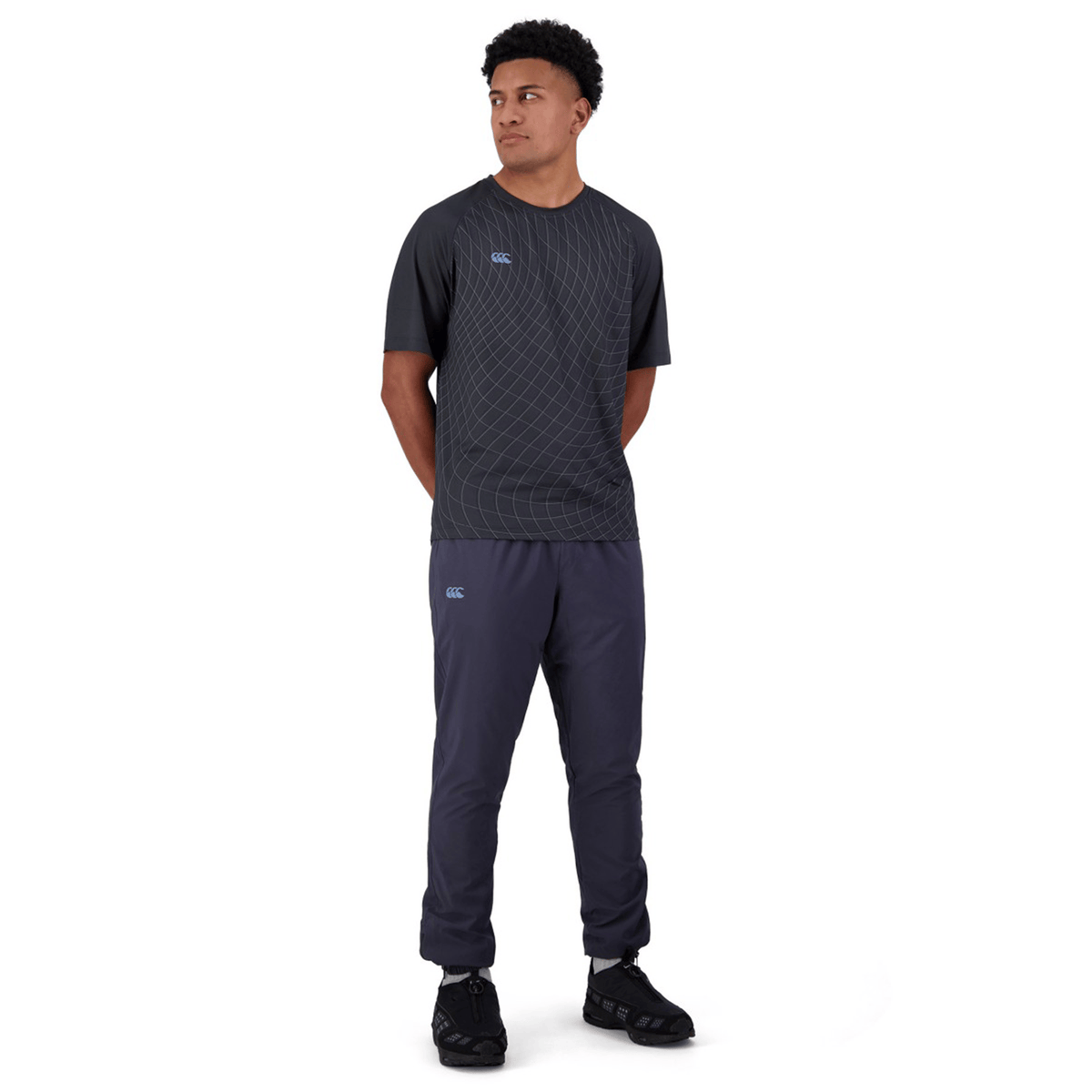 A person dons a dark geometric T-shirt, Canterbury's Tempo+ Training Trackpant in bi-stretch fabric, and black shoes, standing against a white background with hands behind their back, gazing to the right.