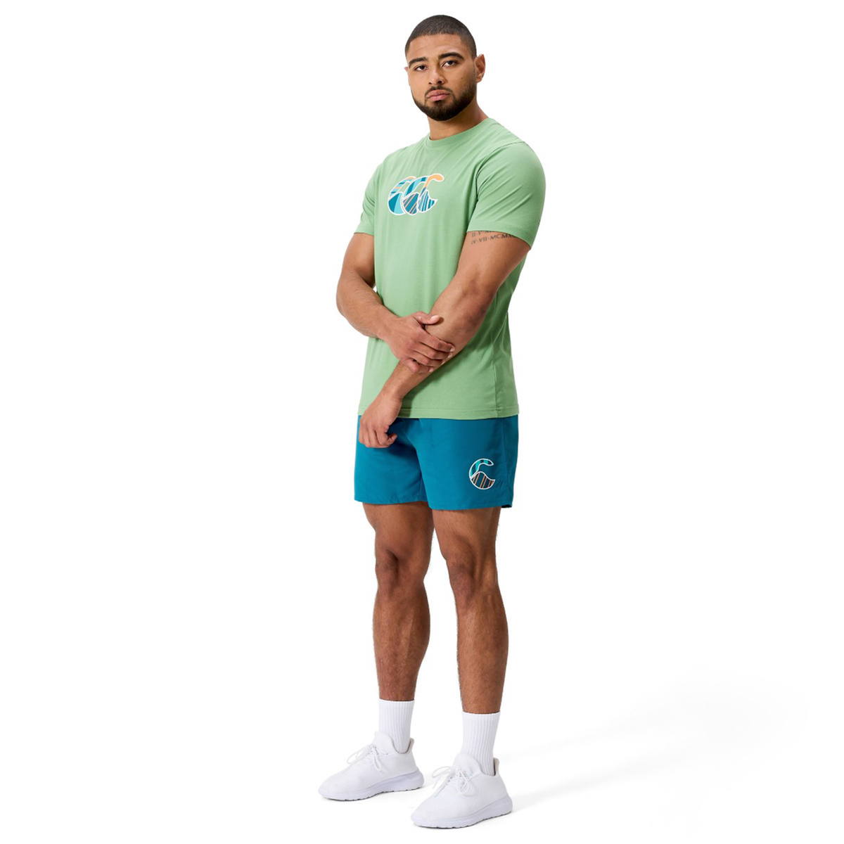 A man stands against a white background wearing a green graphic t-shirt, Canterbury Uglies Logo Tactic Shorts in blue, and white sneakers.
