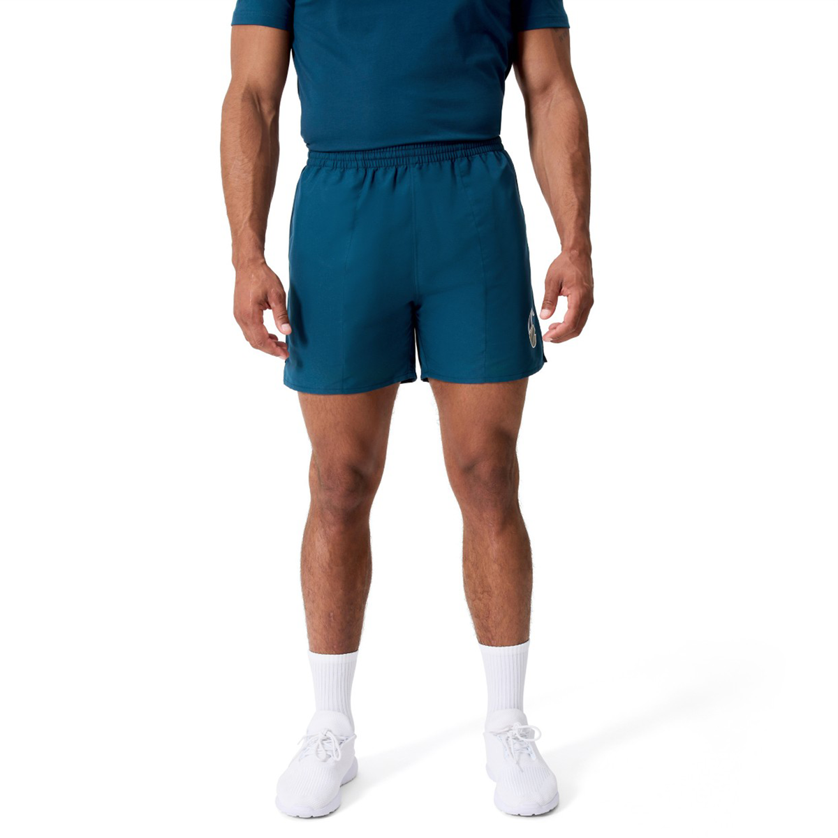 A man wearing a blue t-shirt, Canterbury Uglies Logo Tactic Shorts, white socks, and white sneakers stands against a plain white background.