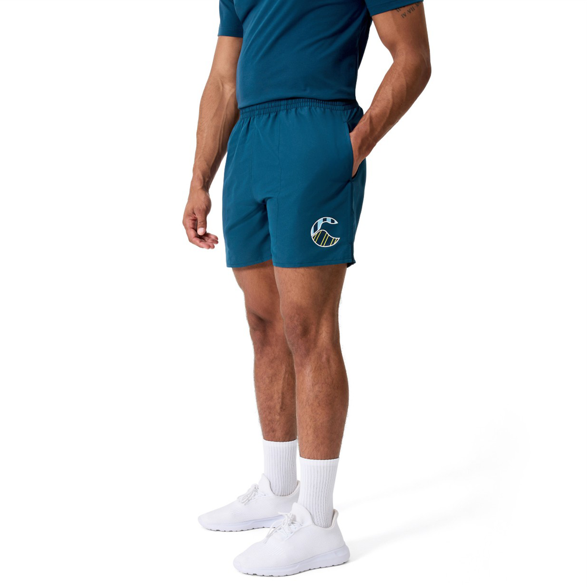 A man wearing Canterbury Uglies Logo Tactic Shorts in teal with the logo on the left leg, a matching t-shirt, white socks, and white sneakers stands against a plain white background.