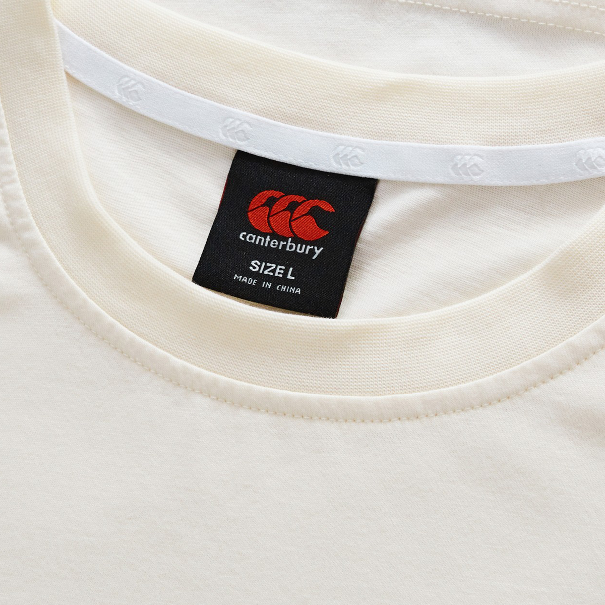 Close-up of the cream-colored Fundamentals CCC Logo T-Shirt by Canterbury, featuring its neckline and a black Canterbury tag marked "Size L, Made in China." A timeless essential from Canterbury.