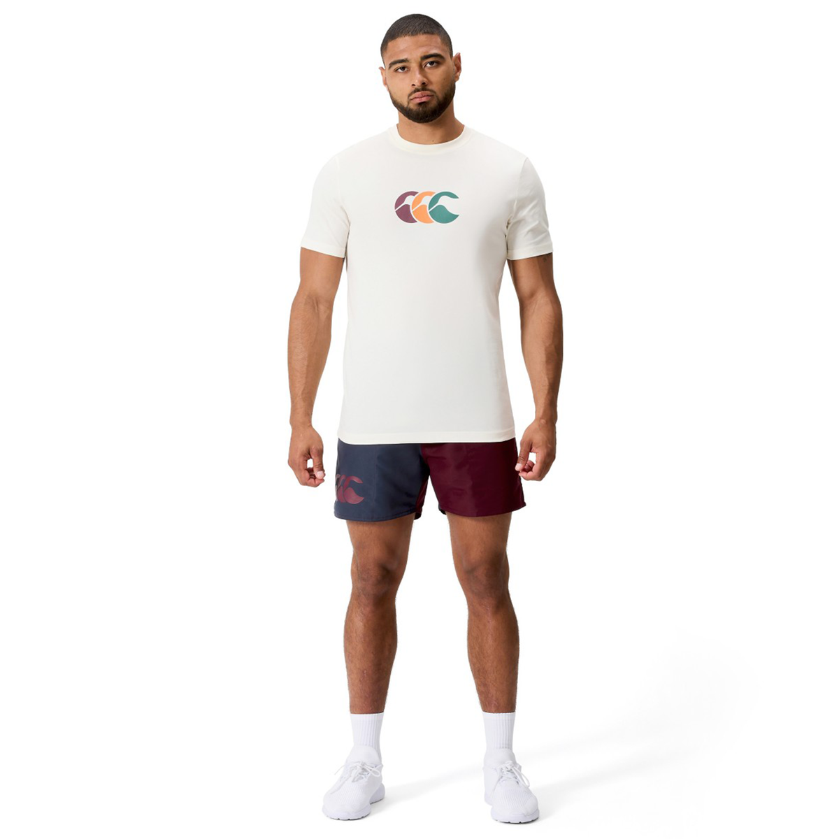 A man faces forward wearing the Canterbury Fundamentals CCC Logo T-Shirt featuring a colorful logo, paired with maroon and navy athletic shorts, white socks, and white sneakers against a plain background.