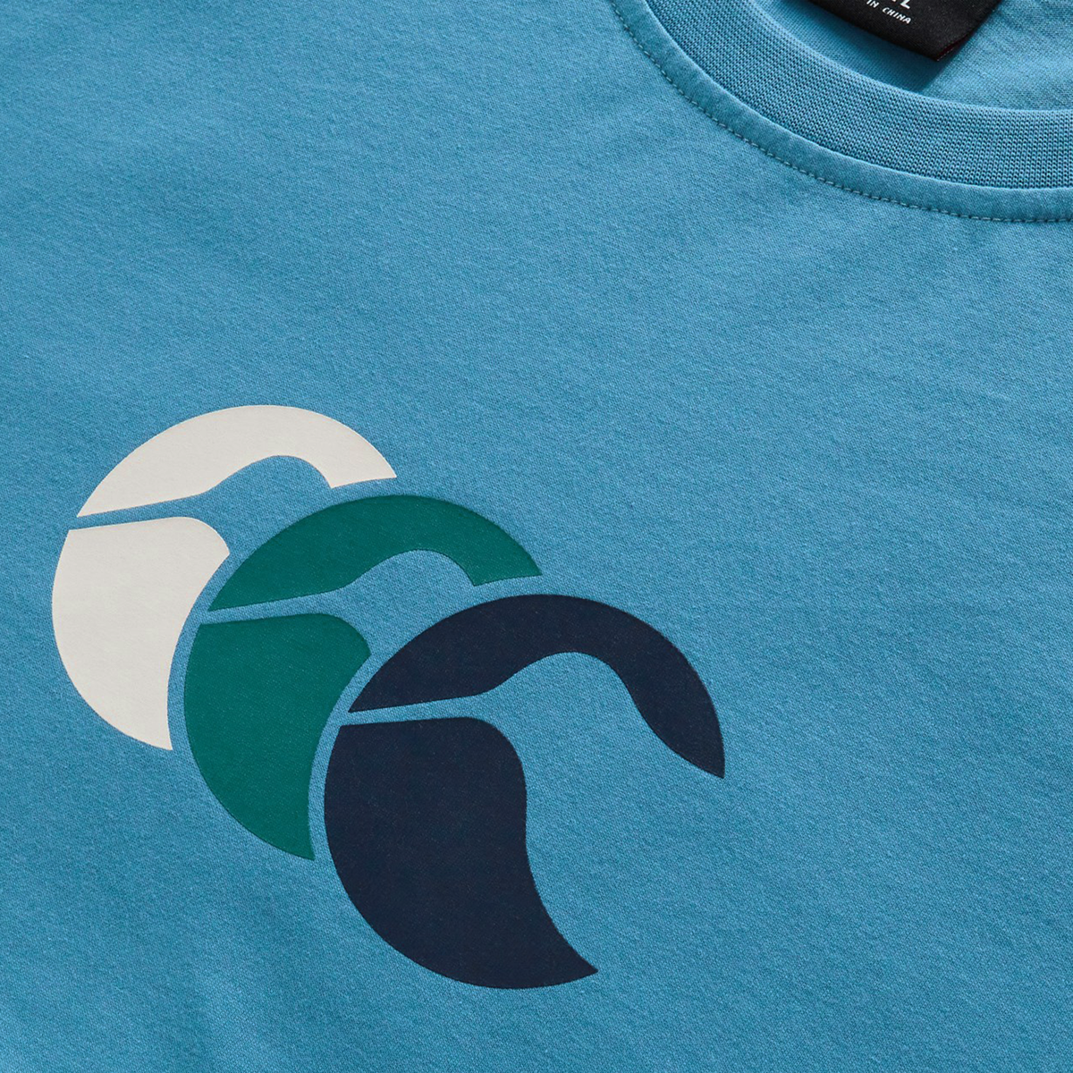 A close-up of the blue Fundamentals CCC Logo T-Shirt by Canterbury, featuring three overlapping white, green, and dark blue wave-like graphics—a stylish nod to the Canterbury brand’s signature Fundamentals collection.