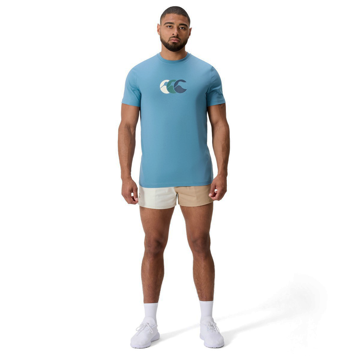 A man stands against a white background in the Canterbury Fundamentals CCC Logo T-Shirt featuring a wave graphic, paired with beige two-tone shorts, white socks, and sneakers—a fresh look for summer style.