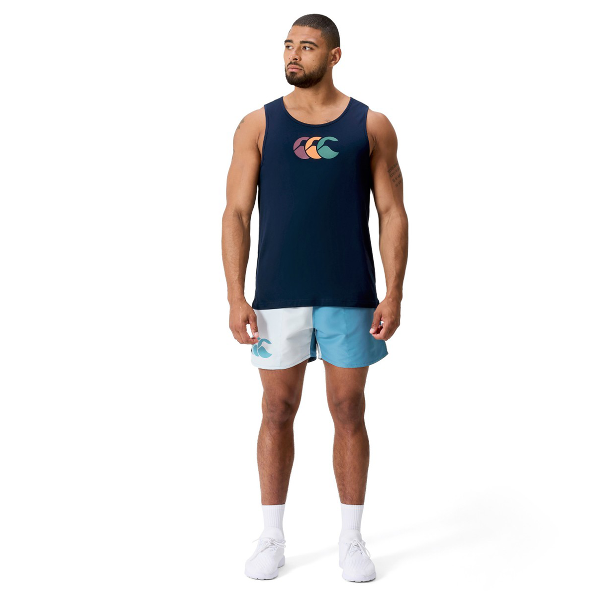 A man wears the Canterbury Fundamentals CCC Logo Tank H225 with a colorful logo, paired with light blue and white athletic shorts and white sneakers, set against a plain white background—a tribute to sporty essentials.