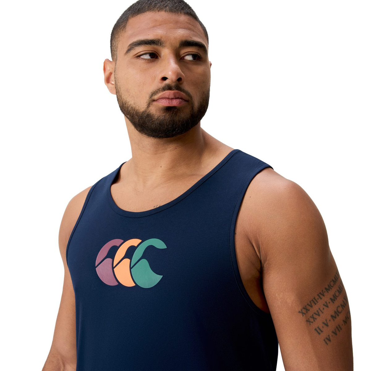 A man in the Canterbury Fundamentals CCC Logo Tank (H225), featuring a multicolored wave logo, looks to his left. He has short hair, a beard, and a Roman numeral tattoo on his upper right arm.