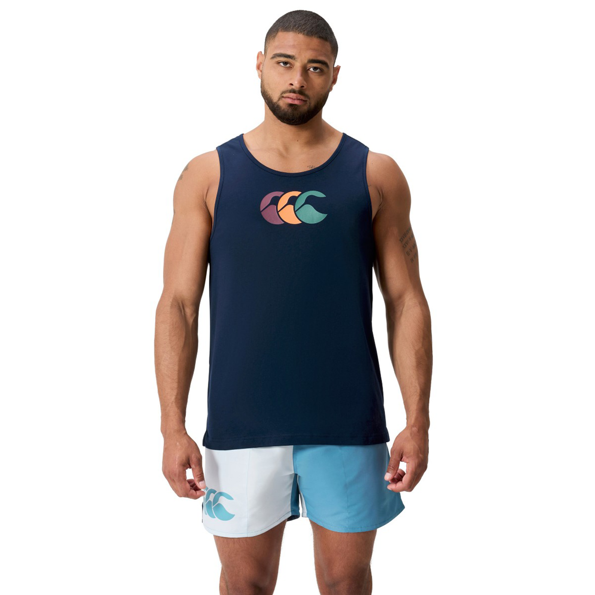 A man faces forward wearing the Canterbury Fundamentals CCC Logo Tank by Canterbury H225 in navy with a colorful logo and light blue and white shorts, highlighting versatile sportswear basics against a plain white background.