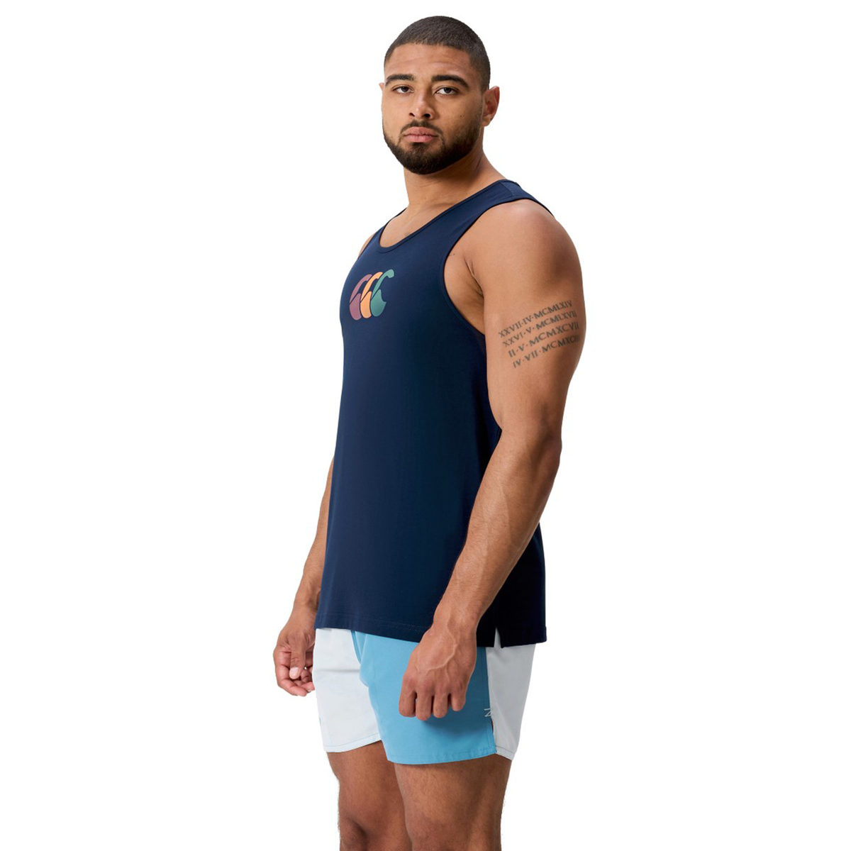 A man stands facing forward against a white background, wearing the Canterbury Fundamentals CCC Logo Tank by Canterbury H225 and light blue shorts, displaying athletic basics with a vibrant logo on his navy tank top.