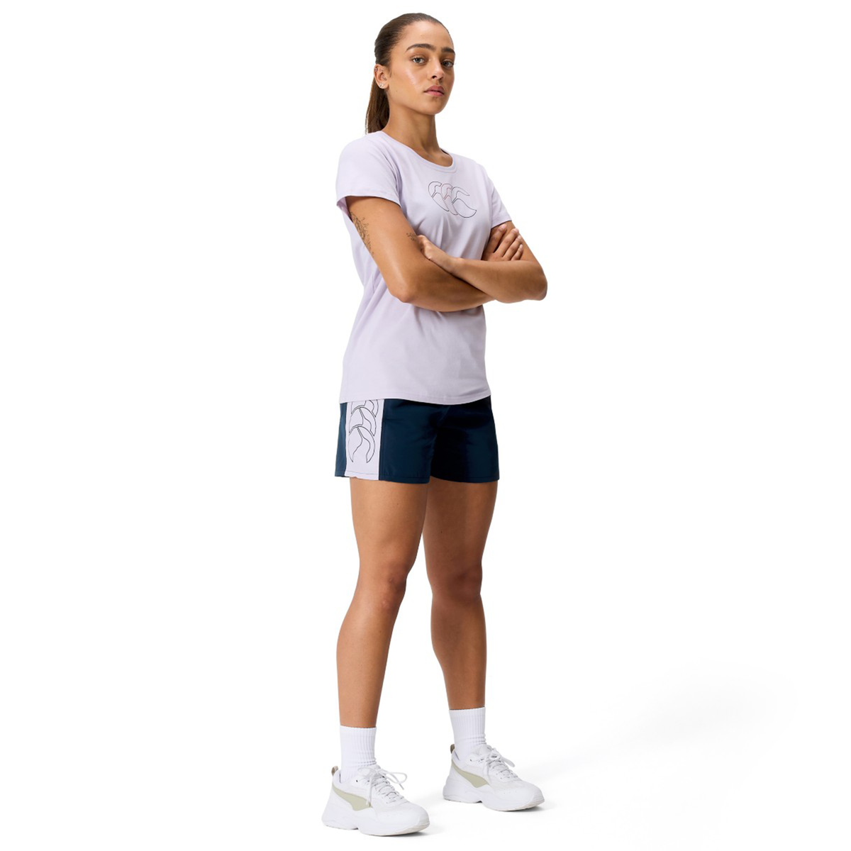 A woman with arms crossed wears a light purple T-shirt, Canterbury Women's Fundamentals 5" Panel Tactic Shorts, white socks, and white sneakers against a plain white background.