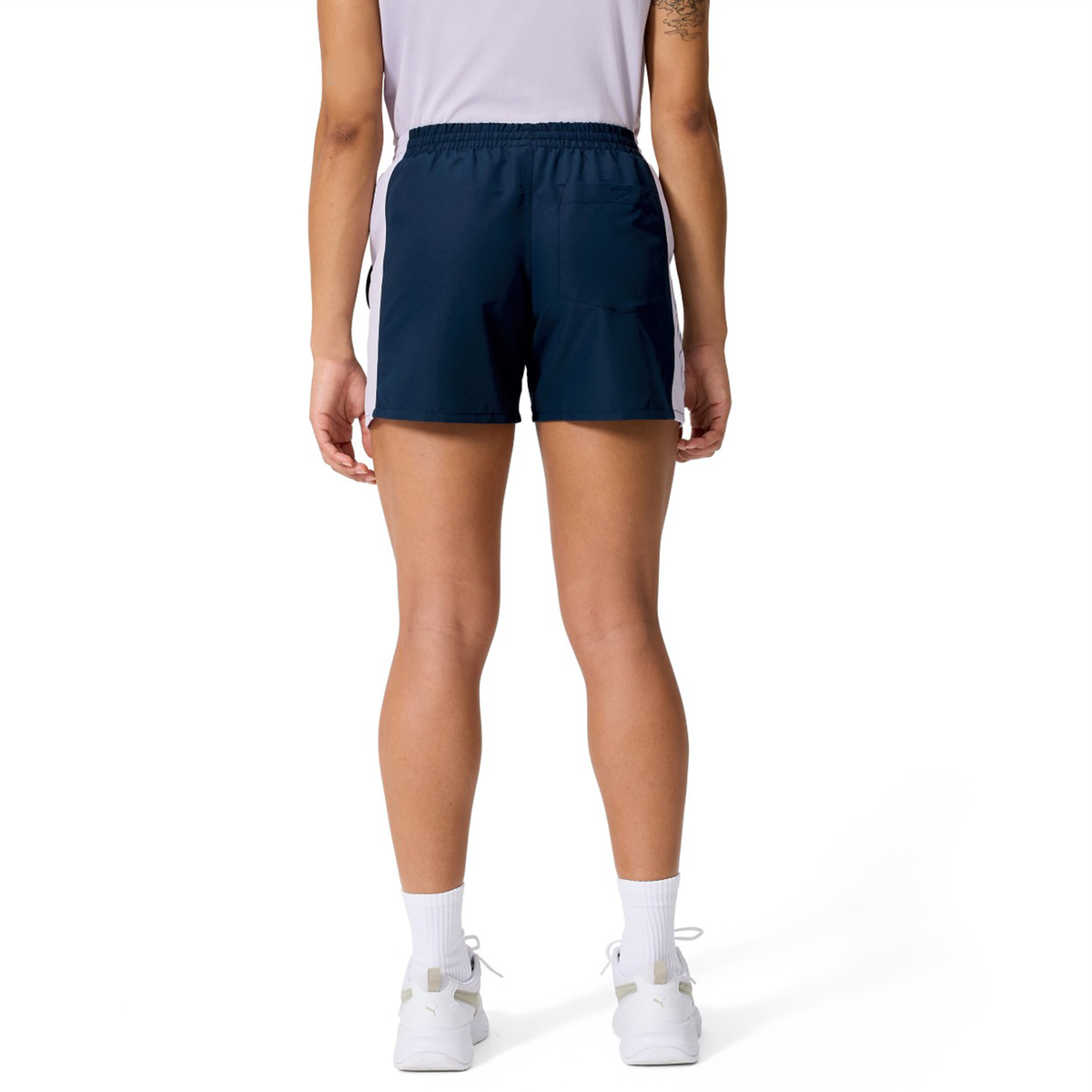 A person stands with their back to the camera, wearing a white shirt and Canterbury Women's Fundamentals 5" Panel Tactic Shorts in navy blue, paired with white socks and sneakers, set against a plain white background.