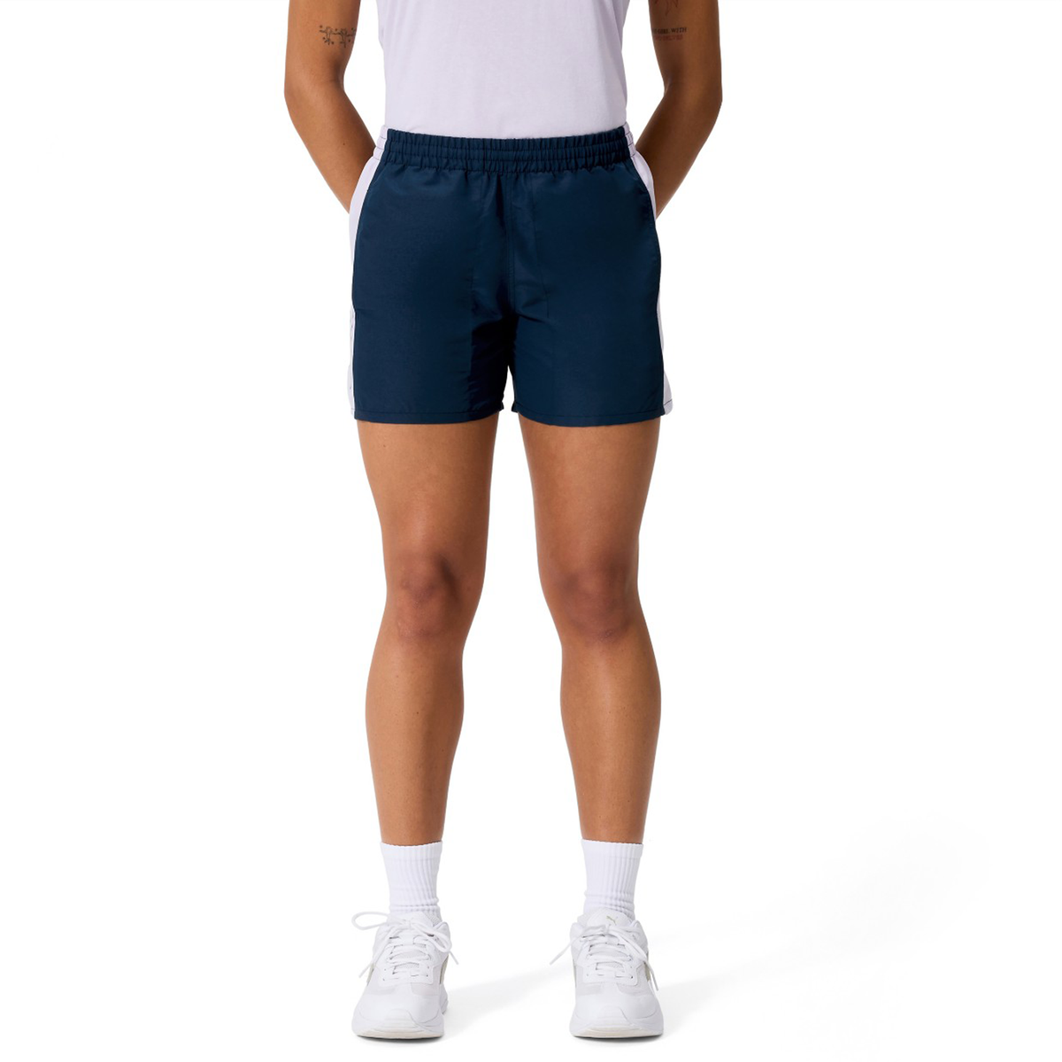 A person stands with hands behind their back, wearing a white t-shirt, Canterbury Women's Fundamentals 5" Panel Tactic Shorts, white socks, and white sneakers against a plain white background.