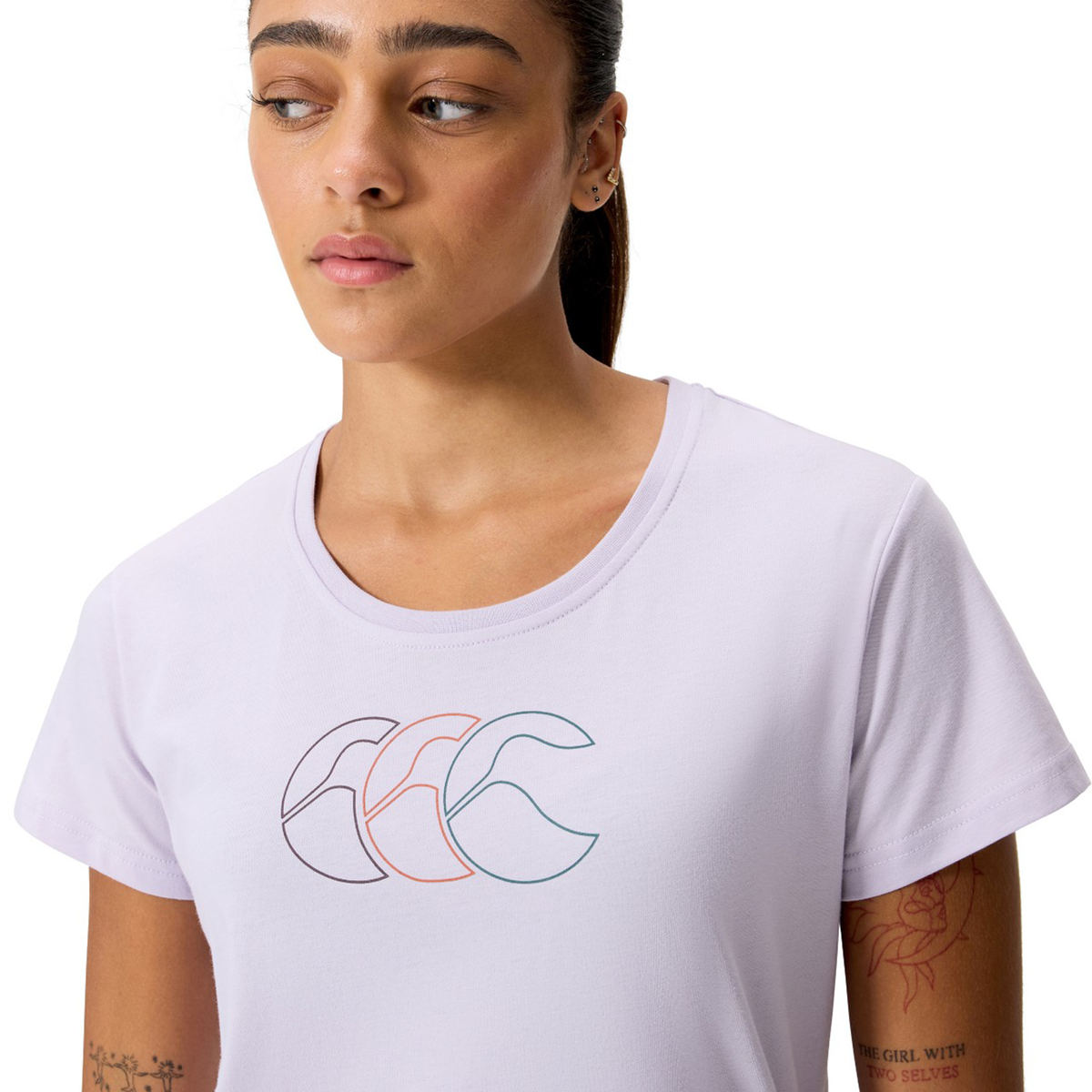 A woman wearing the Women's Fundamentals CCC Logo T-Shirt by Canterbury in light purple, featuring the brand’s minimalist overlapping triple "C" logo on the chest, looks slightly to her left.