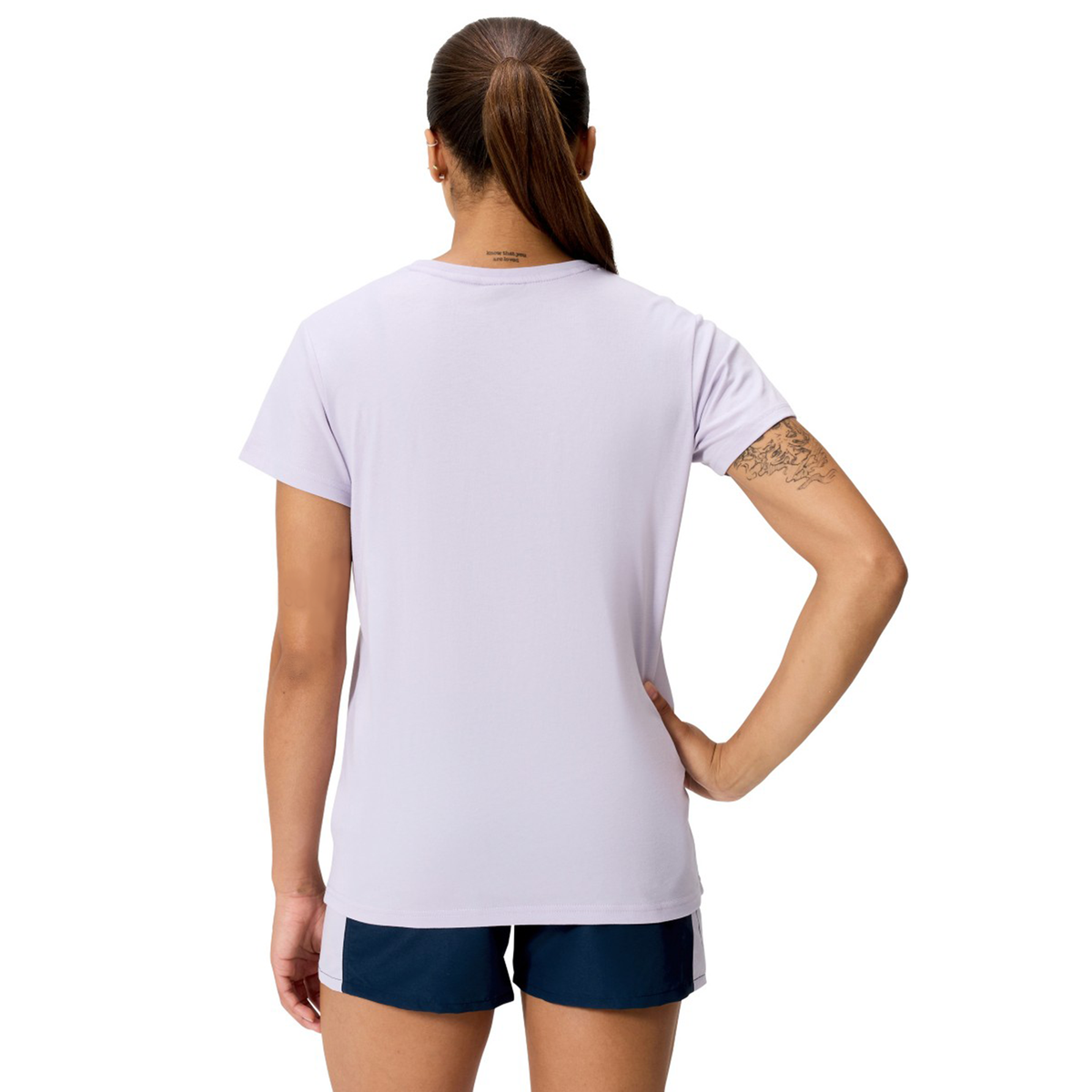 A woman stands facing away, wearing a light purple Women's Fundamentals CCC Logo T-Shirt by Canterbury and navy shorts, with one hand on her hip.