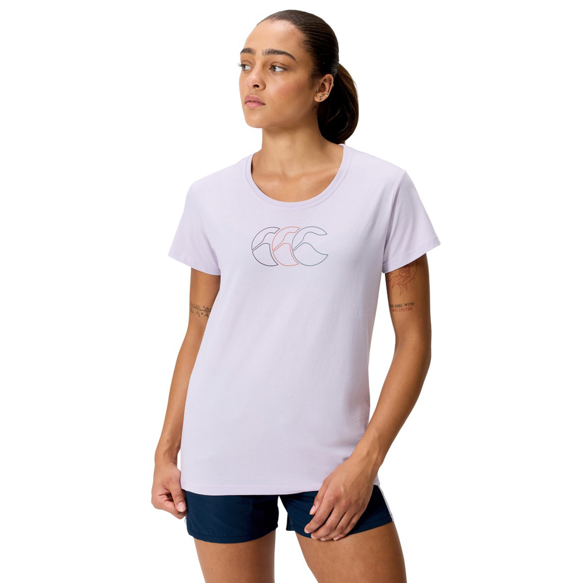 A woman wears the Women's Fundamentals CCC Logo T-Shirt by Canterbury in light purple and navy shorts, standing with her right hand in her pocket and looking to the side.
