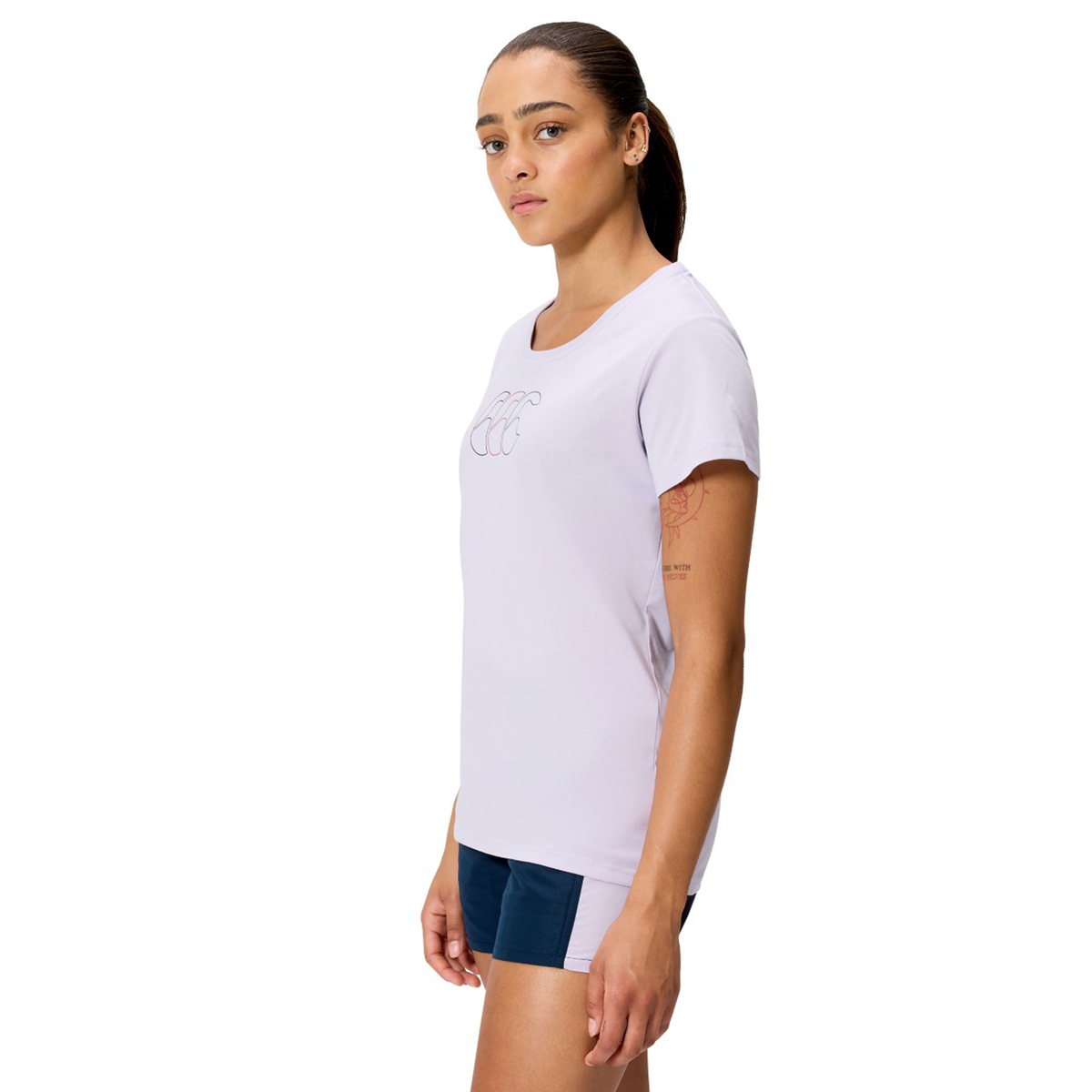 A woman stands in a side pose wearing the Canterbury Women's Fundamentals CCC Logo T-Shirt in light purple and navy blue athletic shorts against a white background.