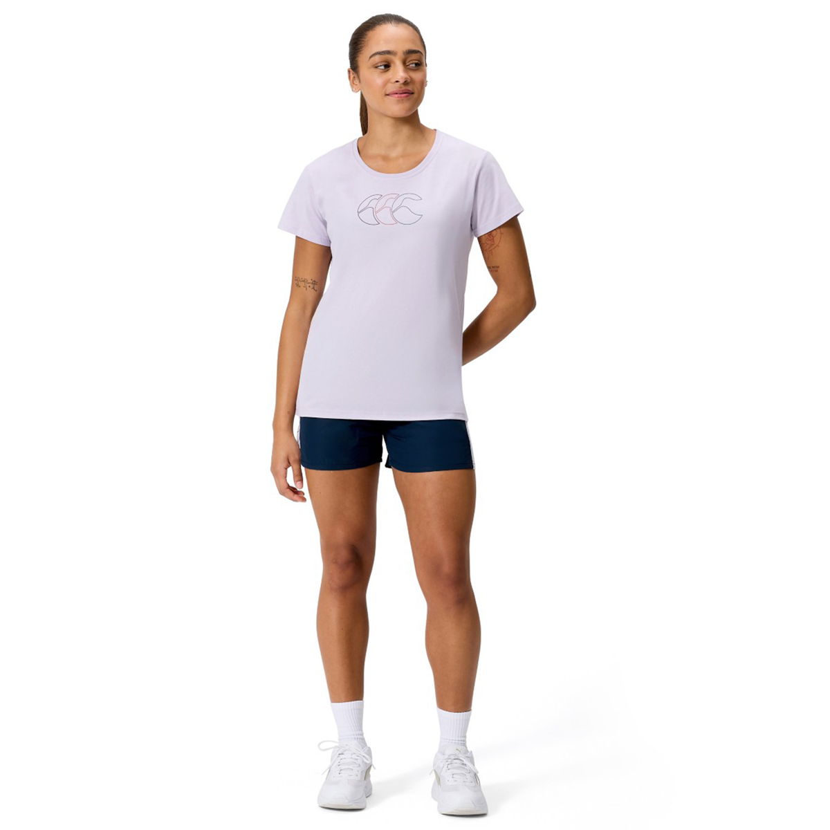 A woman stands facing forward against a white background, wearing a light purple Canterbury Women's Fundamentals CCC Logo T-Shirt, navy blue shorts, white socks, and white sneakers.