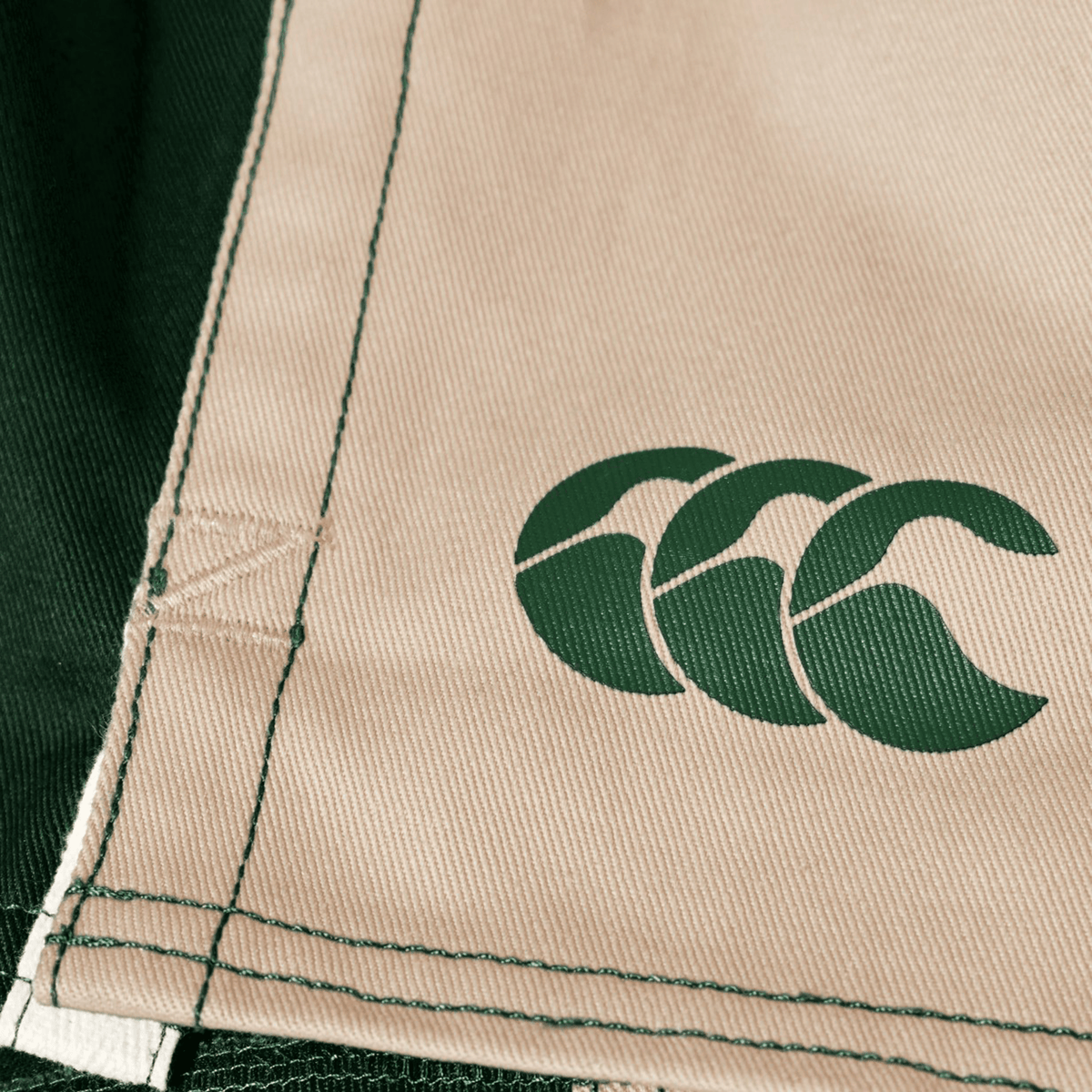 Close-up of beige and dark green Canterbury Fundamental Harlequin Drill Short fabric, featuring visible stitching and a green Canterbury logo.