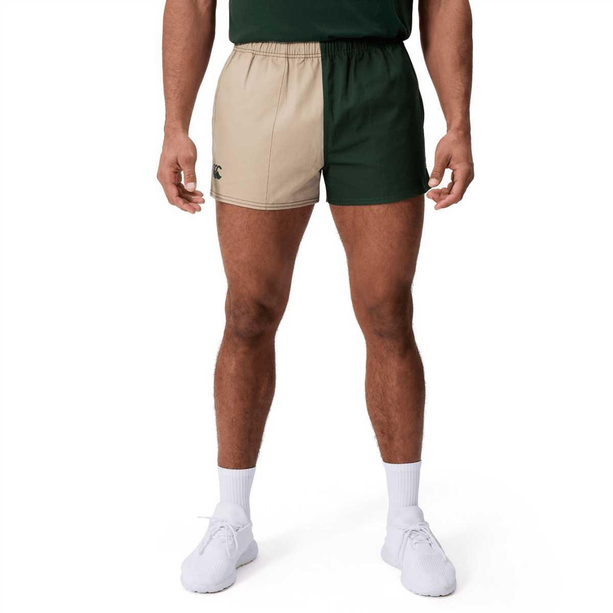 A person wears Canterbury Fundamental Harlequin Drill Shorts by Canterbury—one beige leg, one dark green—with white socks, white shoes, and a green shirt, standing against a white background.