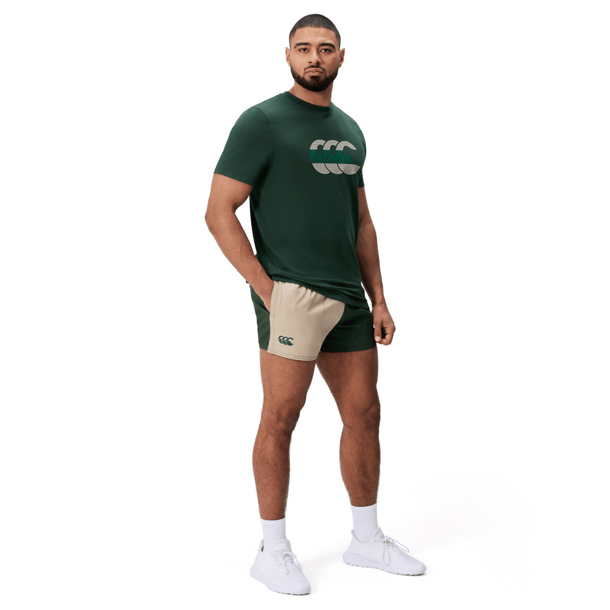 A man stands against a white background wearing a green T-shirt with a logo, Canterbury Fundamental Harlequin Drill Shorts by Canterbury, white socks, and white sneakers.