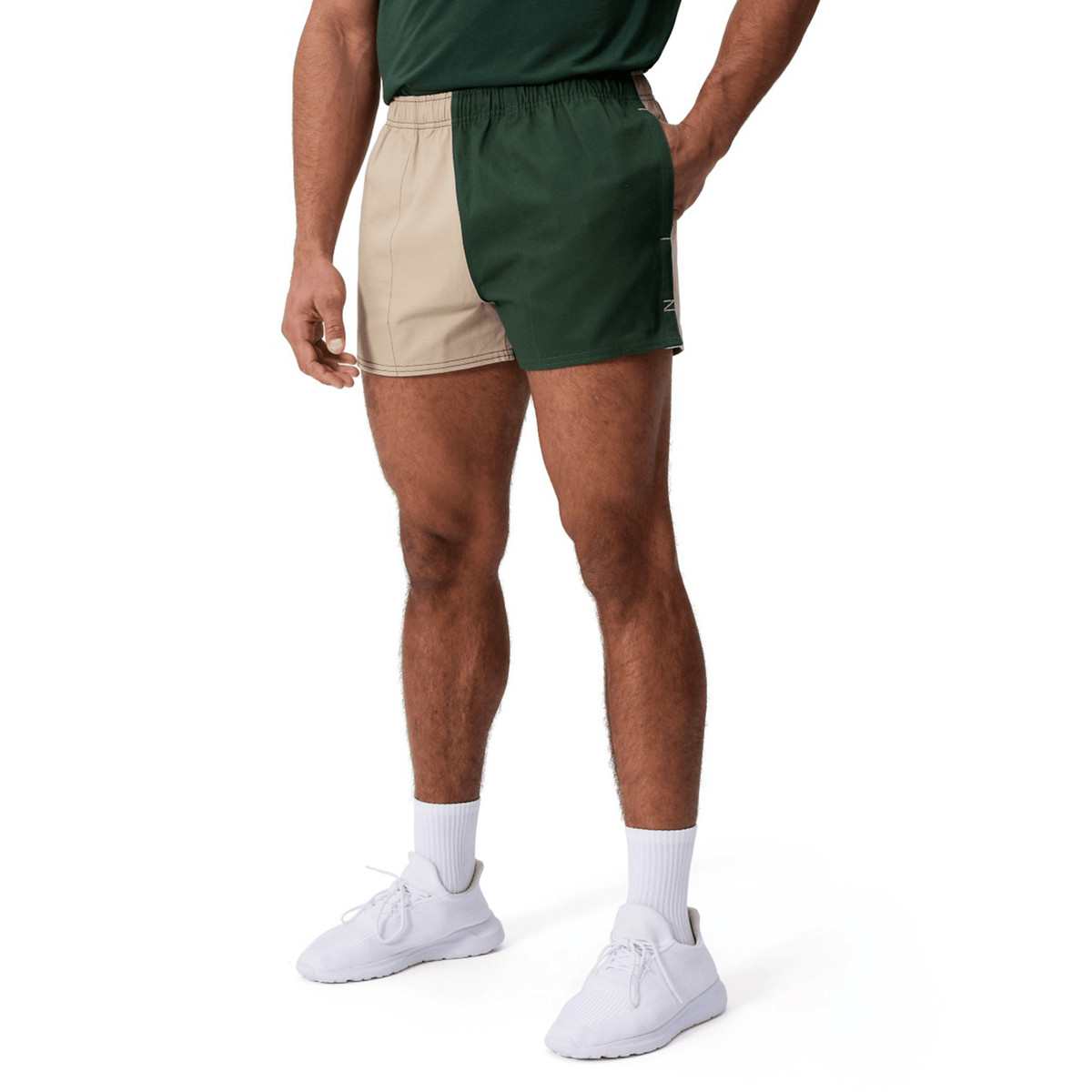A person wears Canterbury Fundamental Harlequin Drill Shorts by Canterbury in green and beige, paired with a green shirt, white socks, and white sneakers, standing against a plain white background.