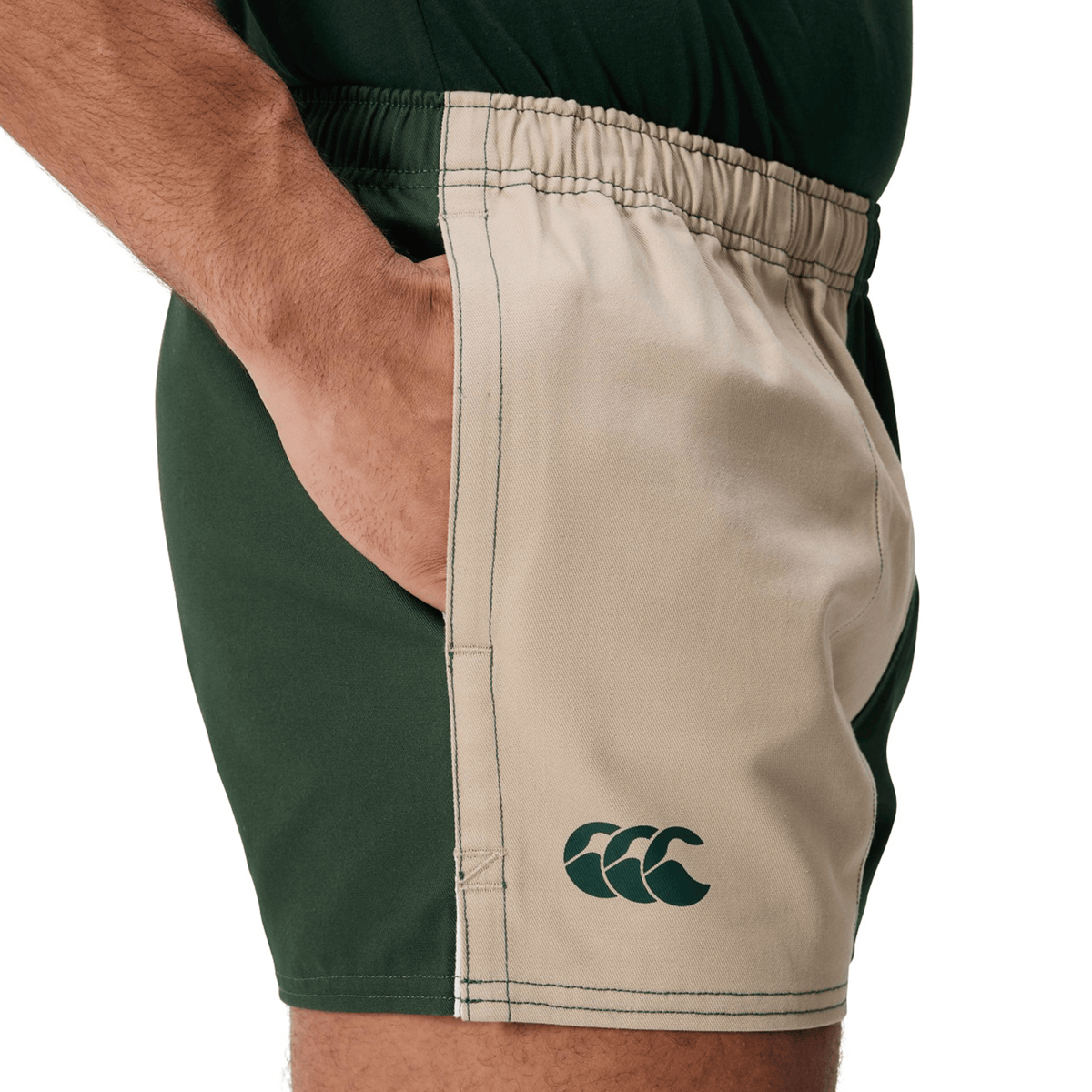 A person wears Canterbury Fundamental Harlequin Drill Shorts by Canterbury, featuring green and beige cotton fabric, a left leg logo, and stands with a hand in the pocket.