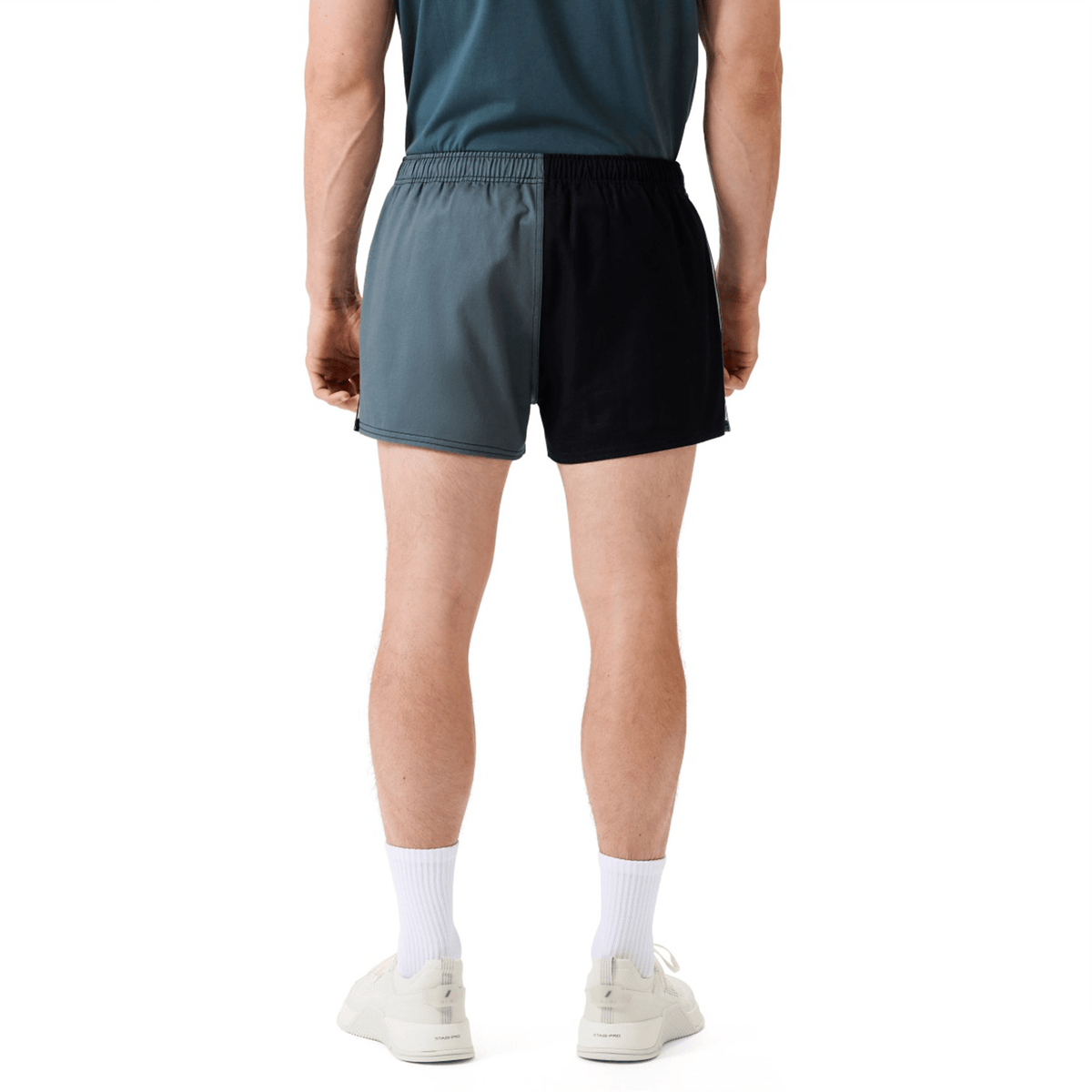 A person stands facing away, wearing a teal shirt, Canterbury Fundamental Harlequin Drill Short by Canterbury in gray and black, white socks, and white sneakers against a plain background.