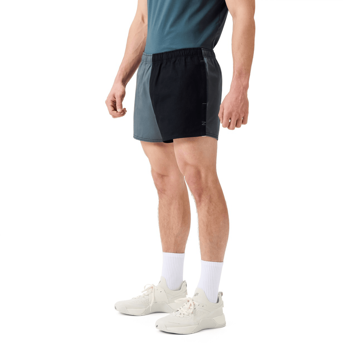 A man wearing a teal shirt, Canterbury Fundamental Harlequin Drill Short by Canterbury, white crew socks, and white sneakers stands against a plain white background.