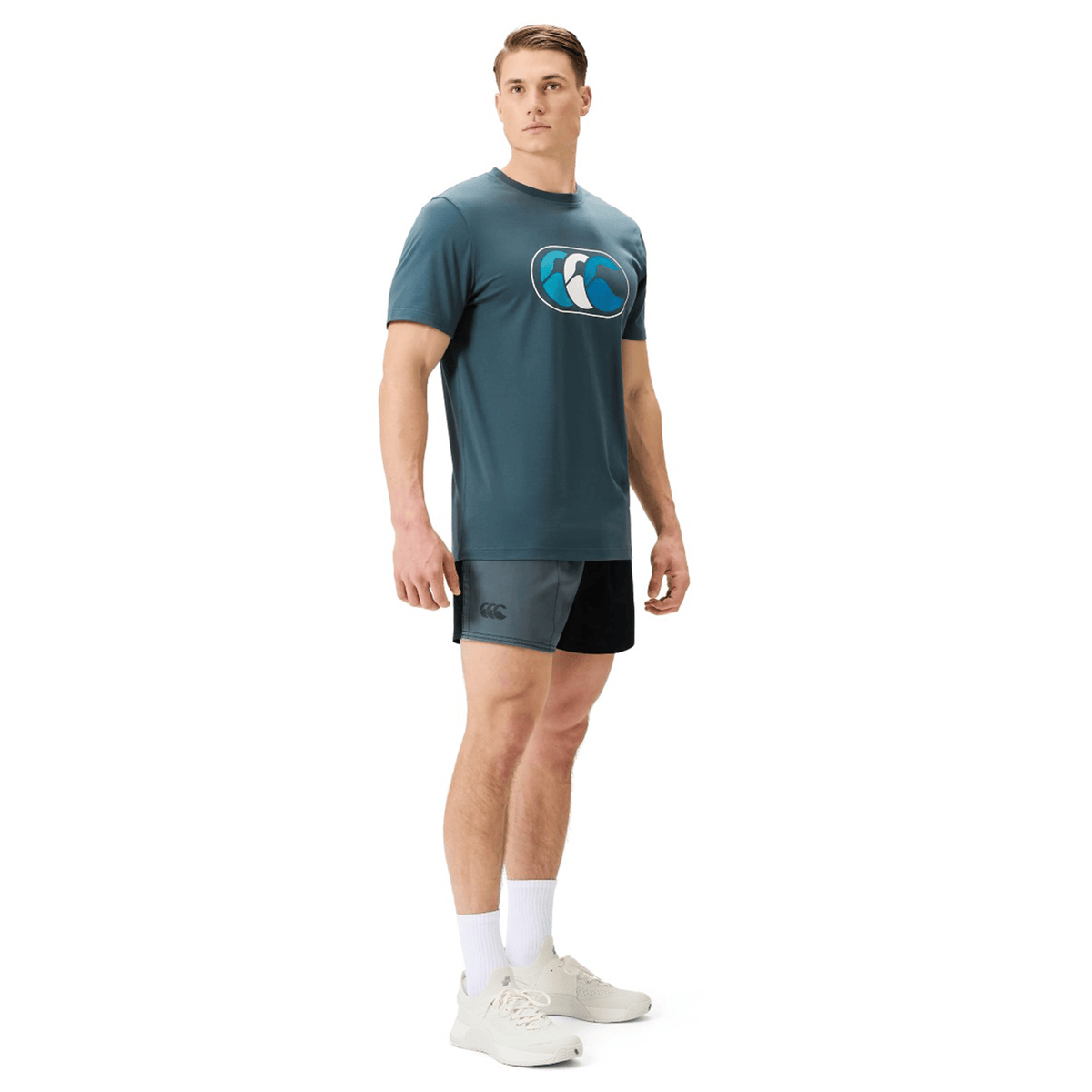 Man standing in an athletic pose, wearing a teal logo t-shirt, Canterbury Fundamental Harlequin Drill Shorts by Canterbury, white socks, and white sneakers against a plain white background.