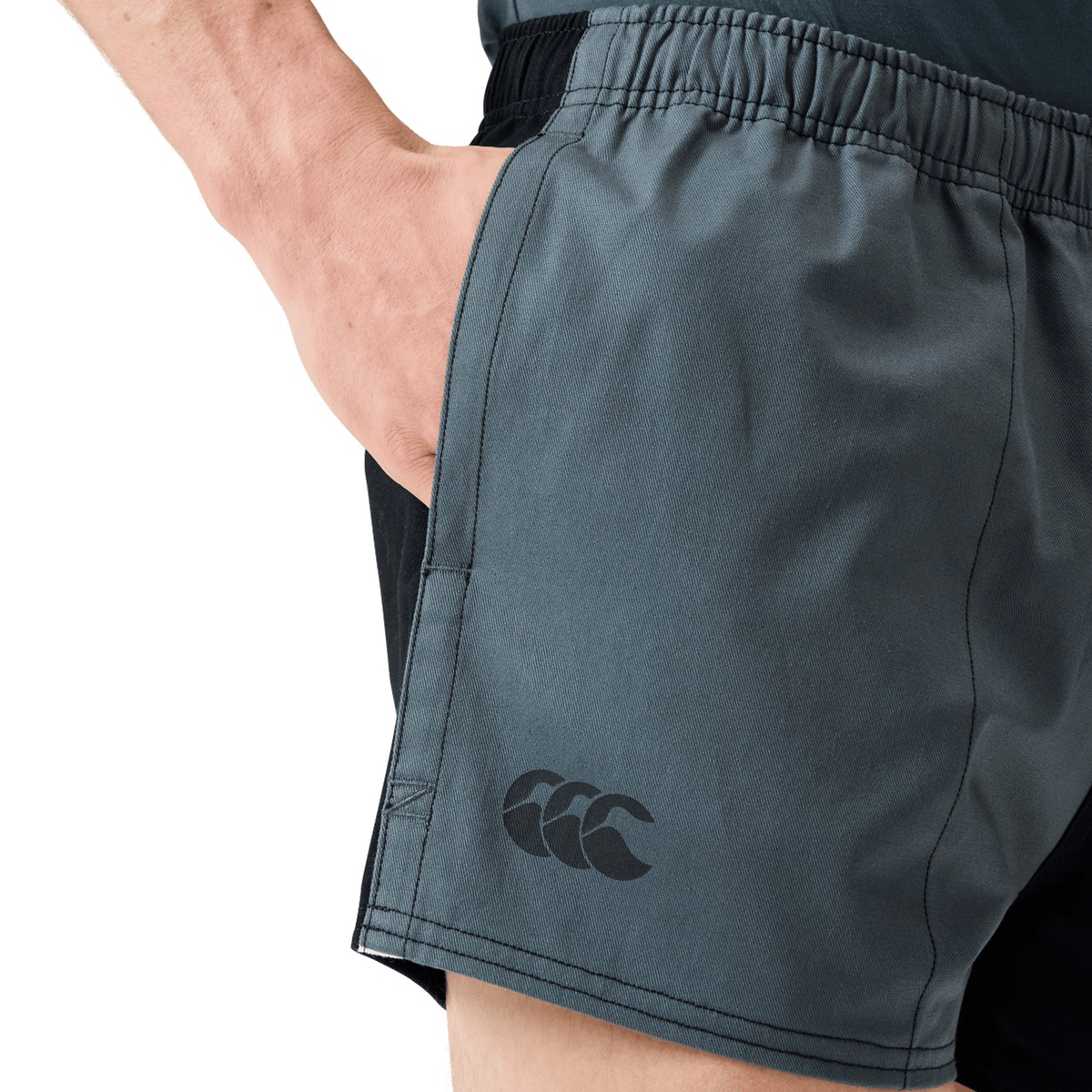 Someone wearing Canterbury Fundamental Harlequin Drill Shorts with a hand in the pocket; these Canterbury shorts have an elastic waistband and a logo visible on the left leg.