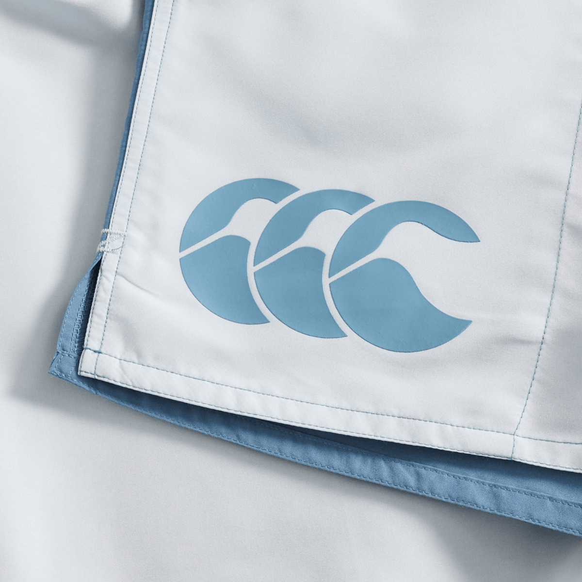 Close-up of light blue and white fabric, characteristic of Canterbury Fundamental Harletic 3″ Rugby Shorts, featuring the Canterbury logo with three overlapping stylized waves or birds printed in blue.