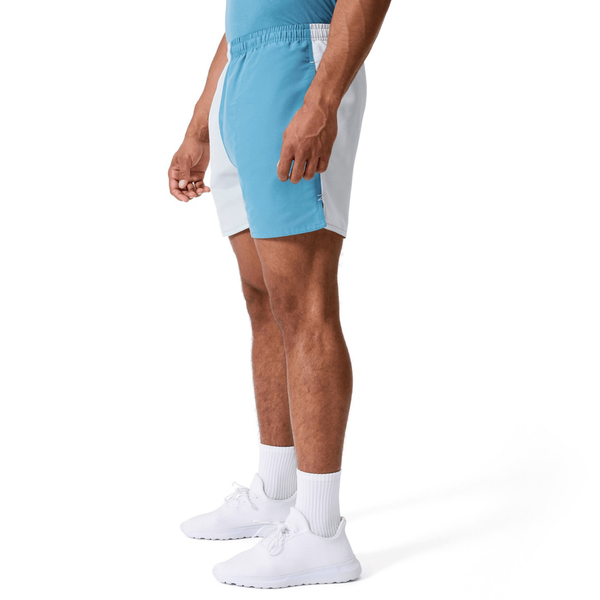 A man stands in profile view against a plain white background wearing Canterbury Fundamental Harletic 3″ Rugby Shorts in light blue and white, paired with a blue shirt, white socks, and white sneakers.