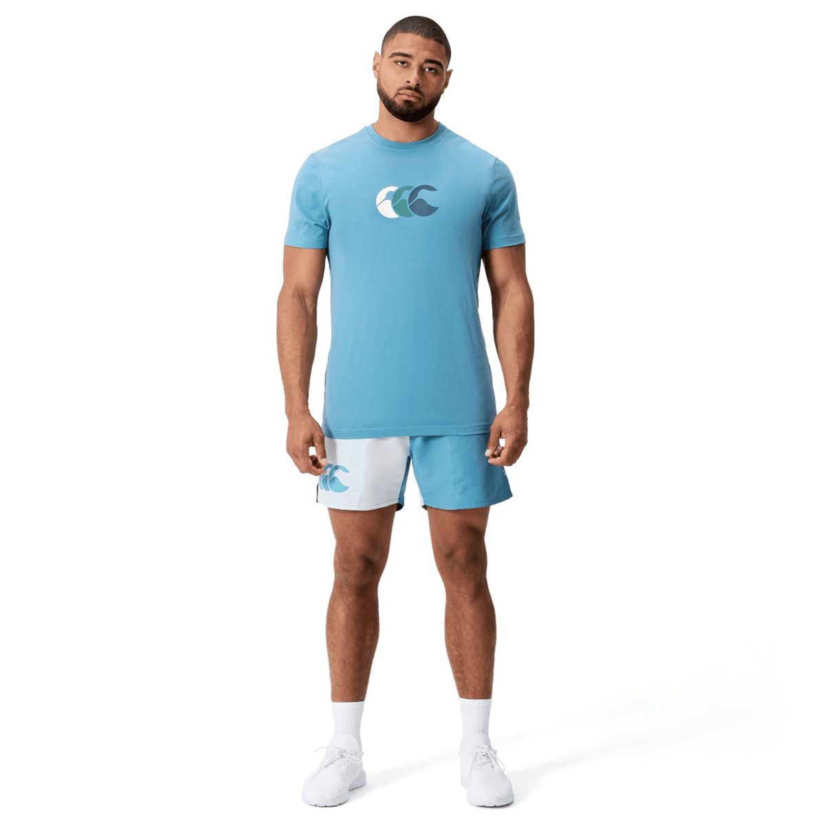 A man stands against a plain white background, wearing a blue t-shirt with a wave logo paired with Canterbury Fundamental Harletic 3″ Rugby Shorts in matching blue and white, along with white socks and shoes.