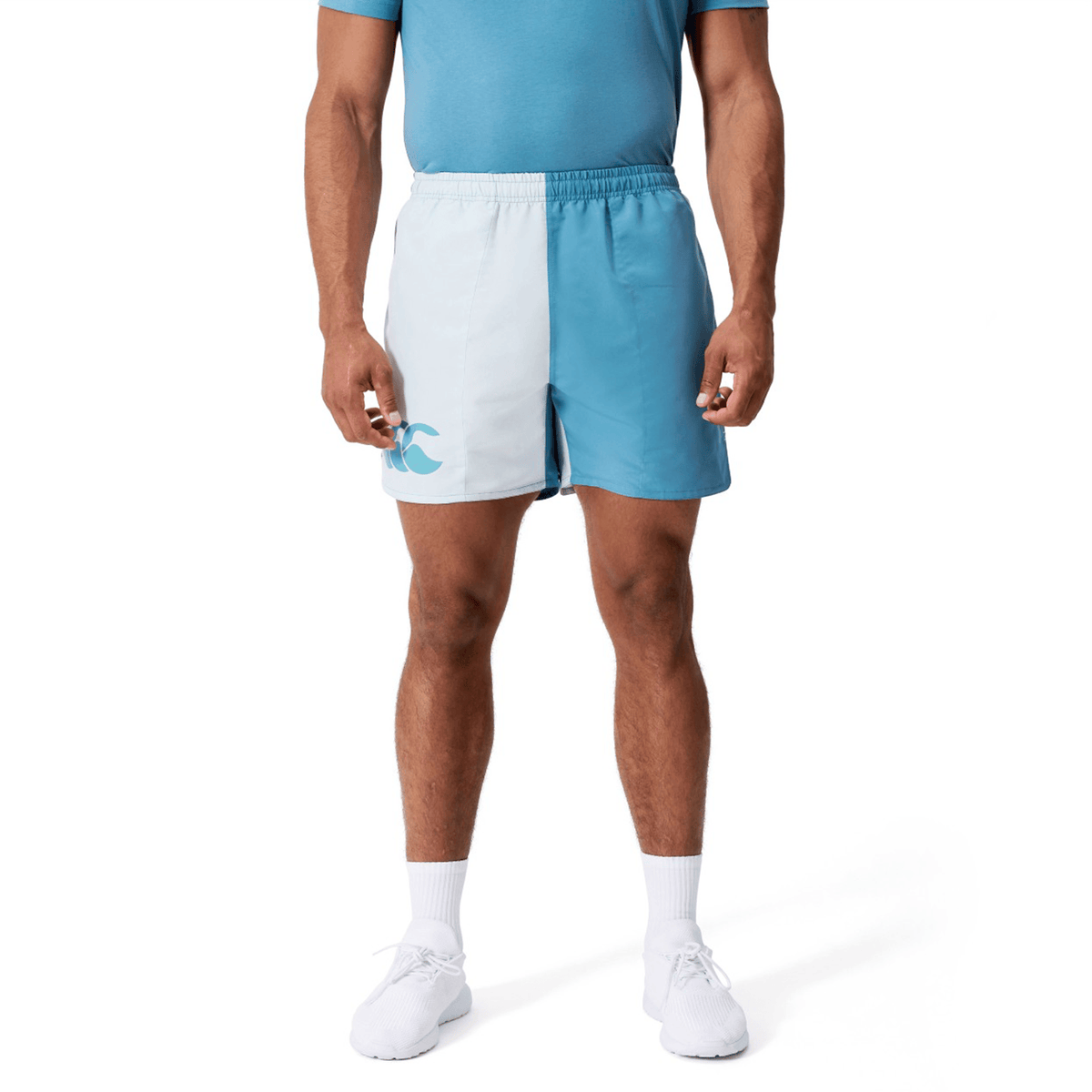 A man wearing Canterbury Fundamental Harletic 3″ Rugby Shorts in light blue and white, a blue t-shirt, white socks, and white shoes stands against a plain white background.