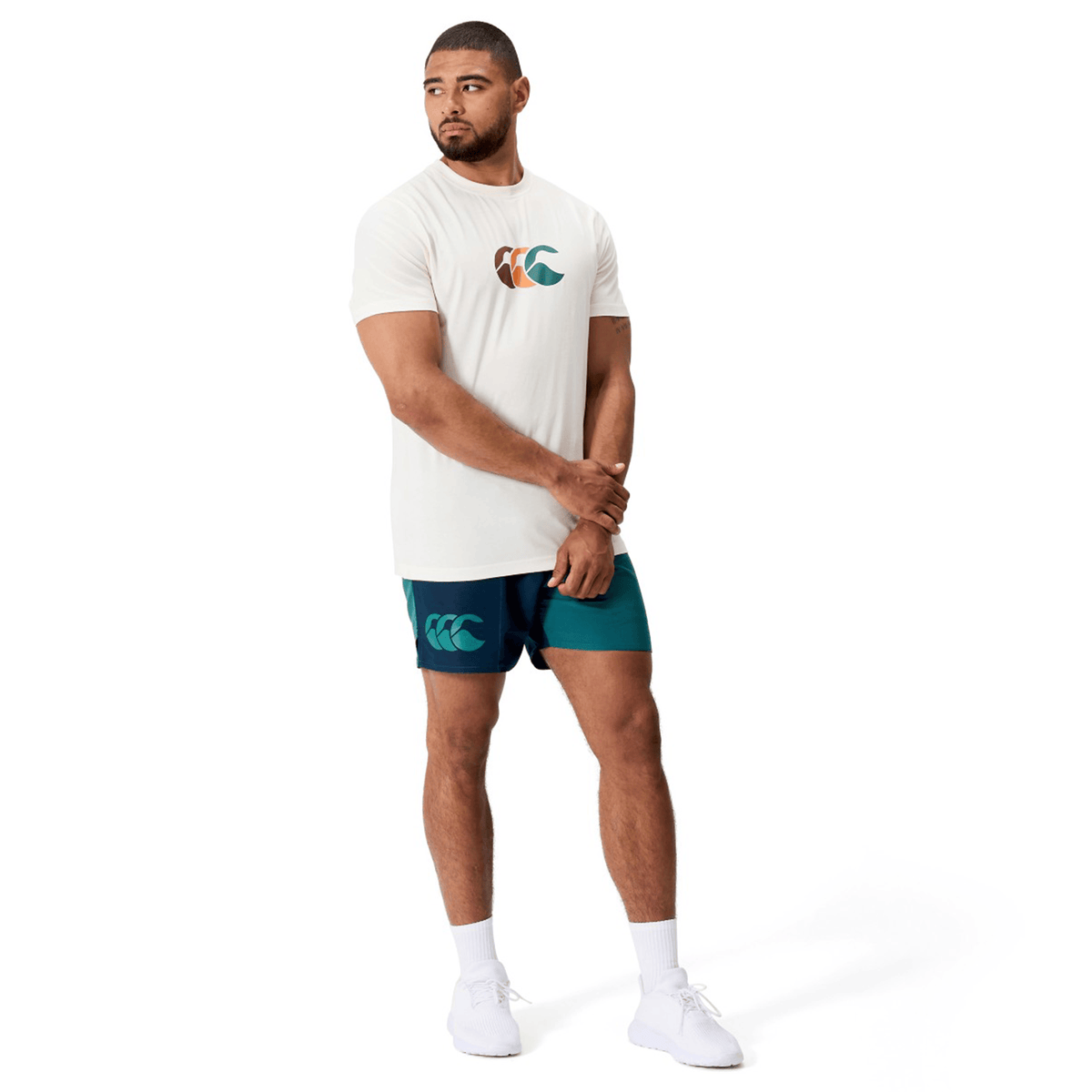 A man wearing a white graphic T-shirt, Canterbury Fundamental Harletic 3″ Rugby Shorts in teal and navy, white socks, and white sneakers stands against a white background.