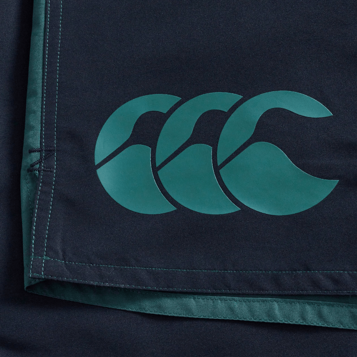 Close-up of Canterbury Fundamental Harletic 3″ Rugby Shorts in dark fabric, showcasing a teal overlapping triple-curved Canterbury logo on the lower edge, visible stitching detail, and a comfortable 3-inch inseam.